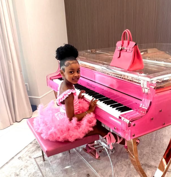 A young child sits at a transparent pink piano, wearing a pink ruffled tutu dress. A pink handbag is placed on the piano