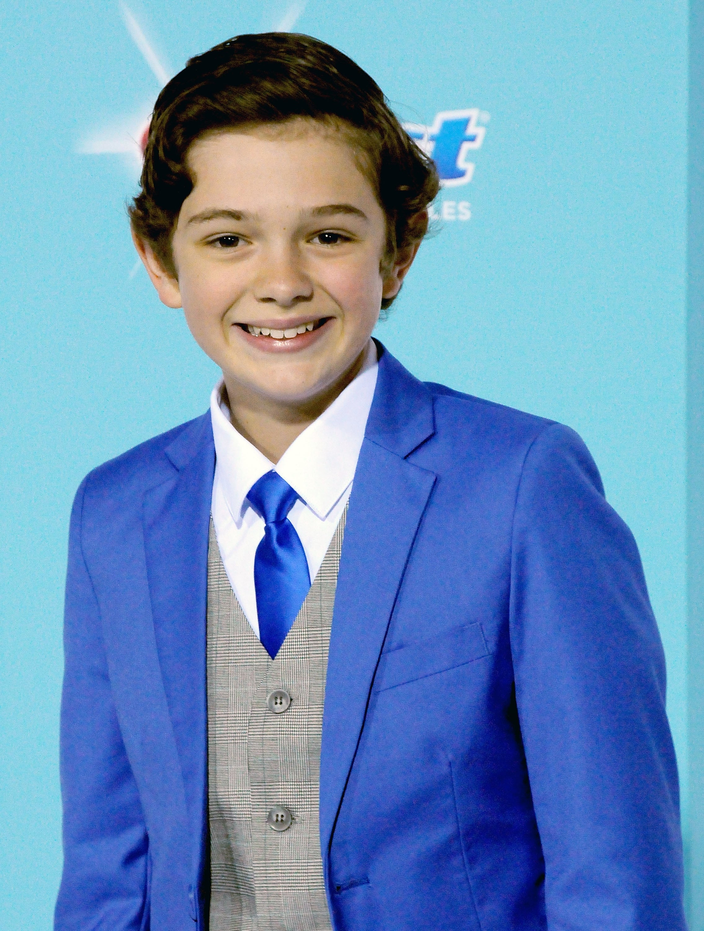 Young person at an event, wearing a blue suit with a gray vest and blue tie, smiling at the camera against a light background