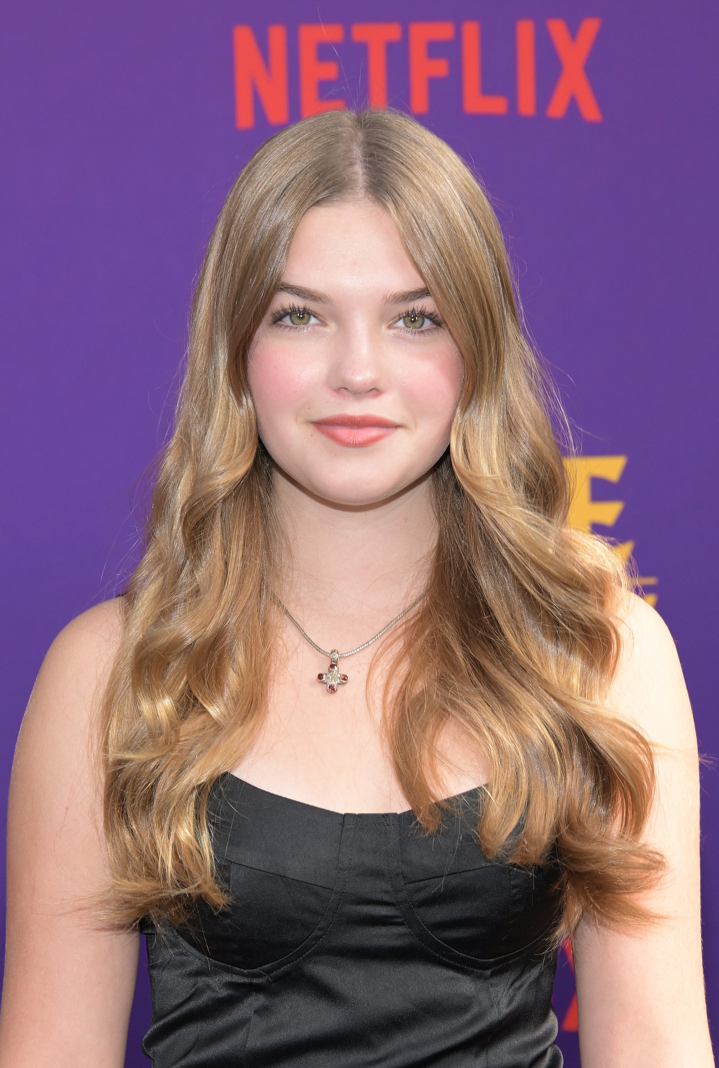 Person with long wavy hair in a sleeveless black dress at a Netflix event. Purple backdrop with Netflix logo visible