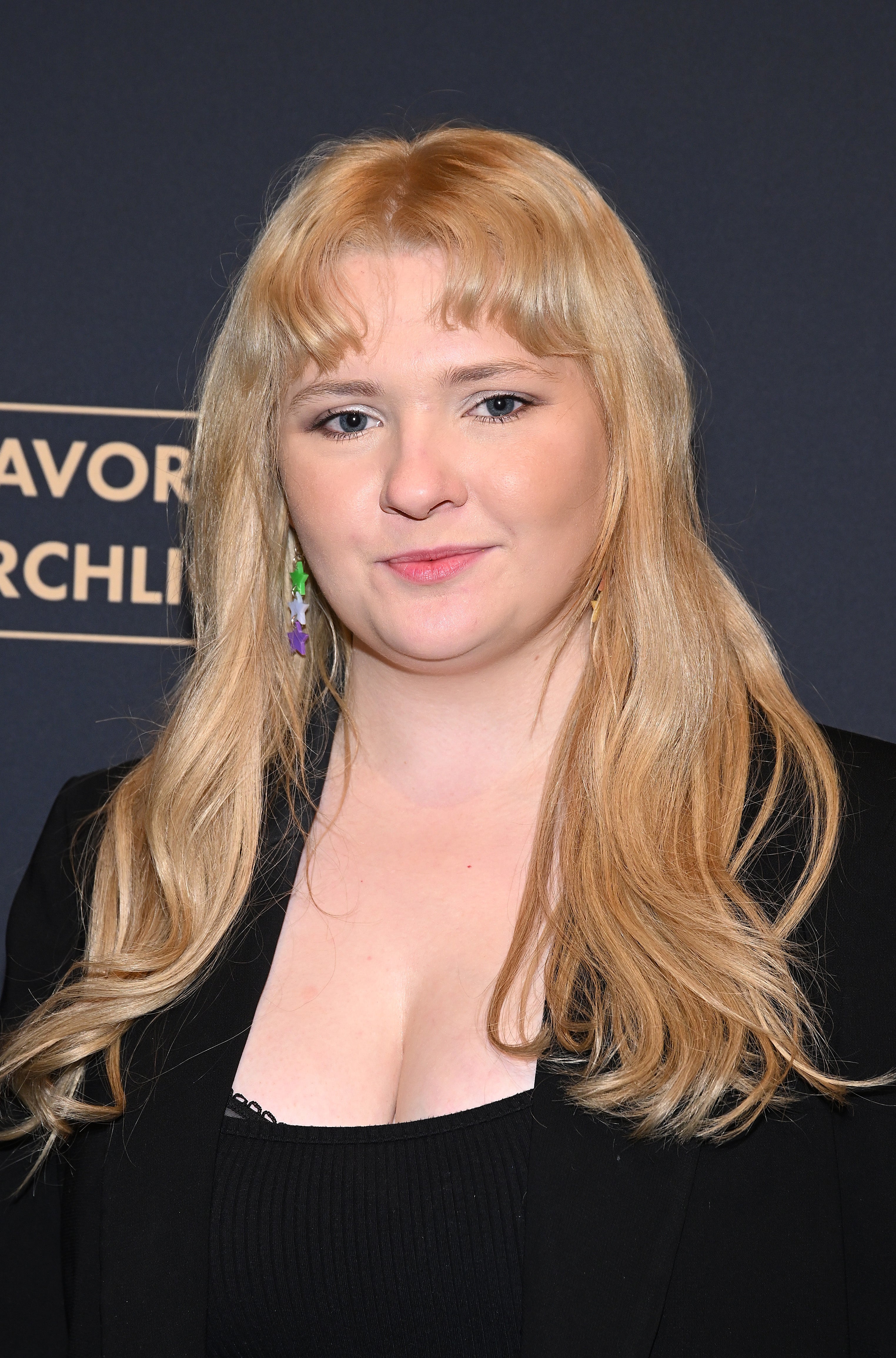 Person with long hair wearing a black blazer at a formal event backdrop