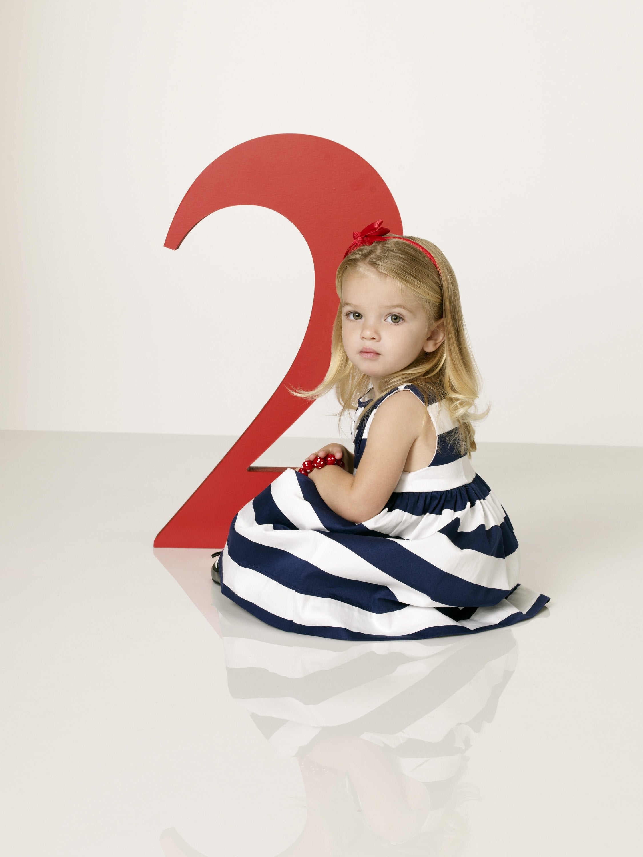 A young child in a striped dress sits beside a large number 2, looking at the camera