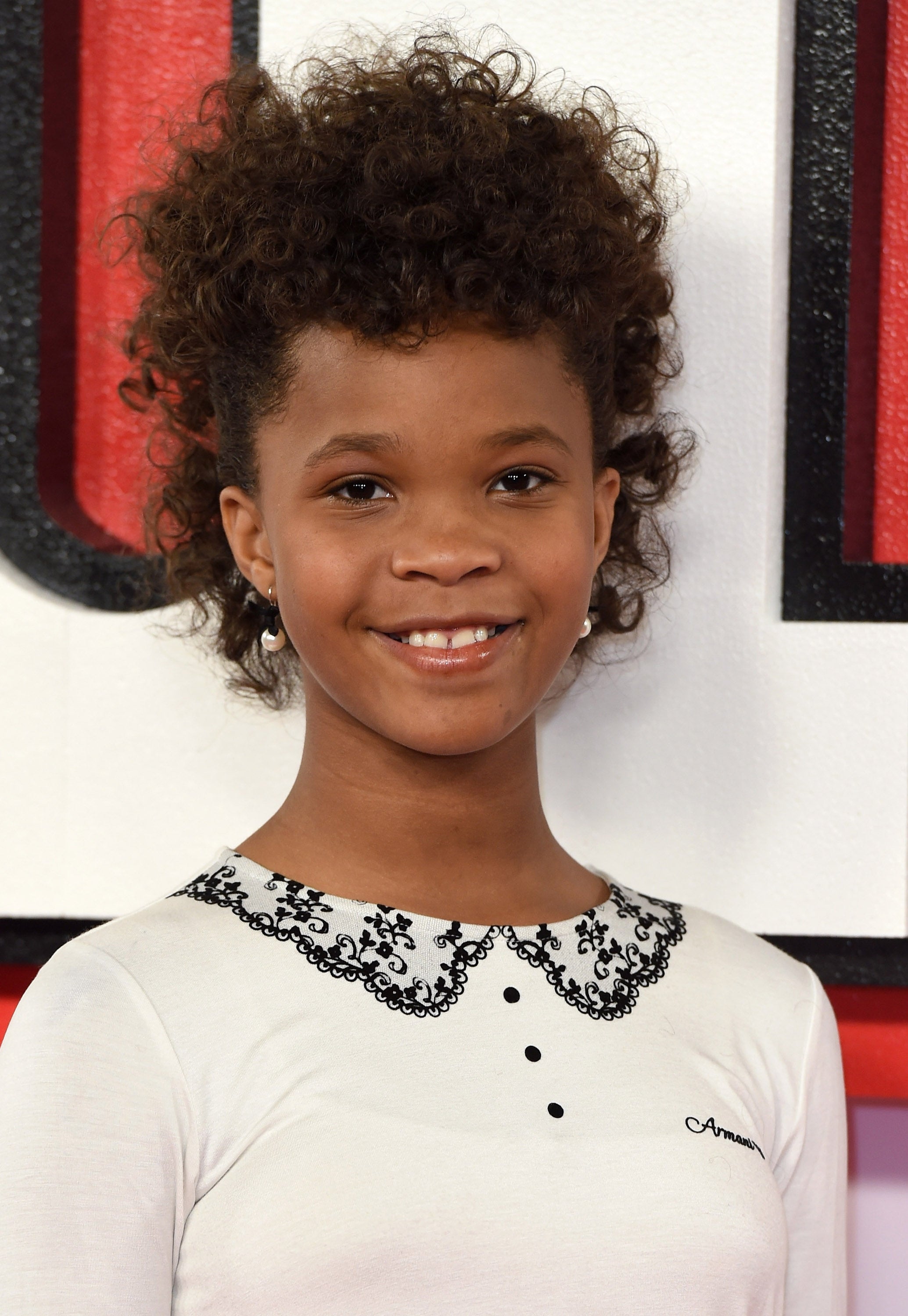 Young girl with curly hair smiles, wearing a white top with a lace collar and small buttons, at a formal event