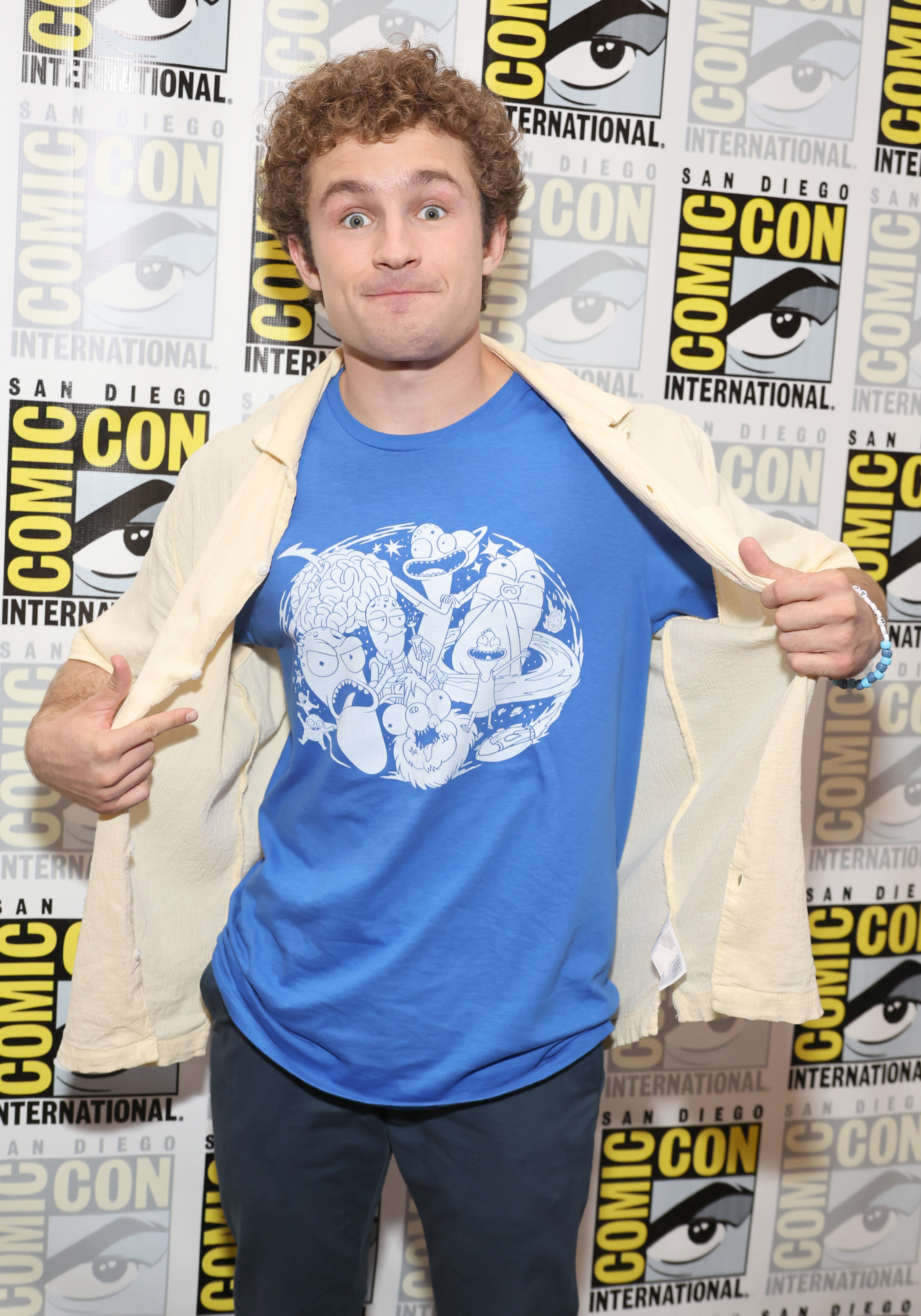Person in casual outfit, open beige cardigan, blue graphic t-shirt, making a surprised face at a Comic-Con event. Comic-Con logo backdrop