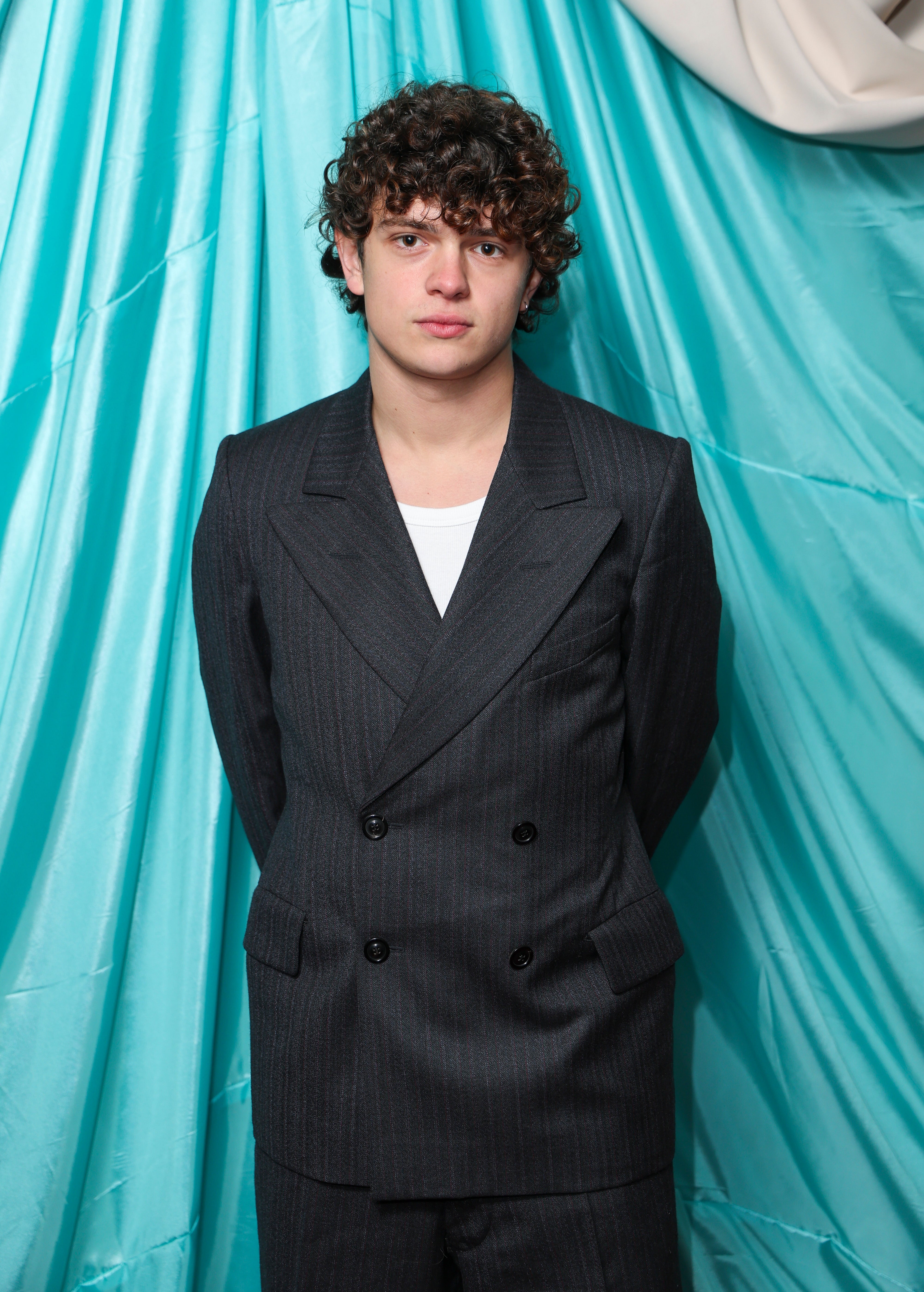Person in a tailored double-breasted suit stands against a draped backdrop, posing with hands behind their back