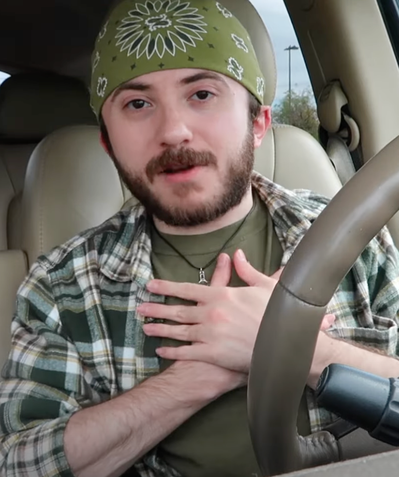 Person in car wearing a bandana and plaid shirt, gesturing with hands over chest, speaking to camera