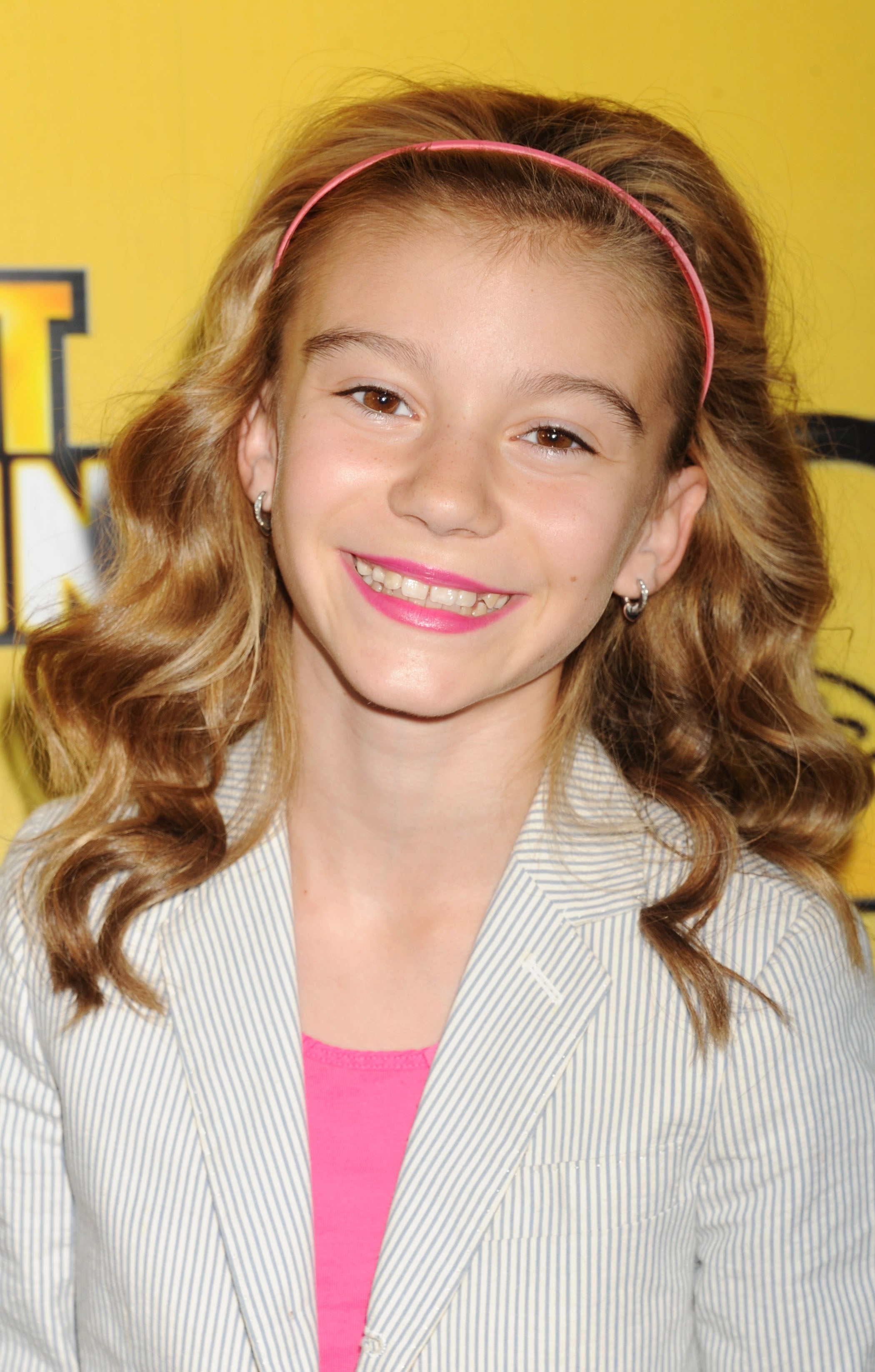 Young girl smiling, wearing a pinstripe blazer and pink top with a headband, at a public event. Background features out-of-focus text