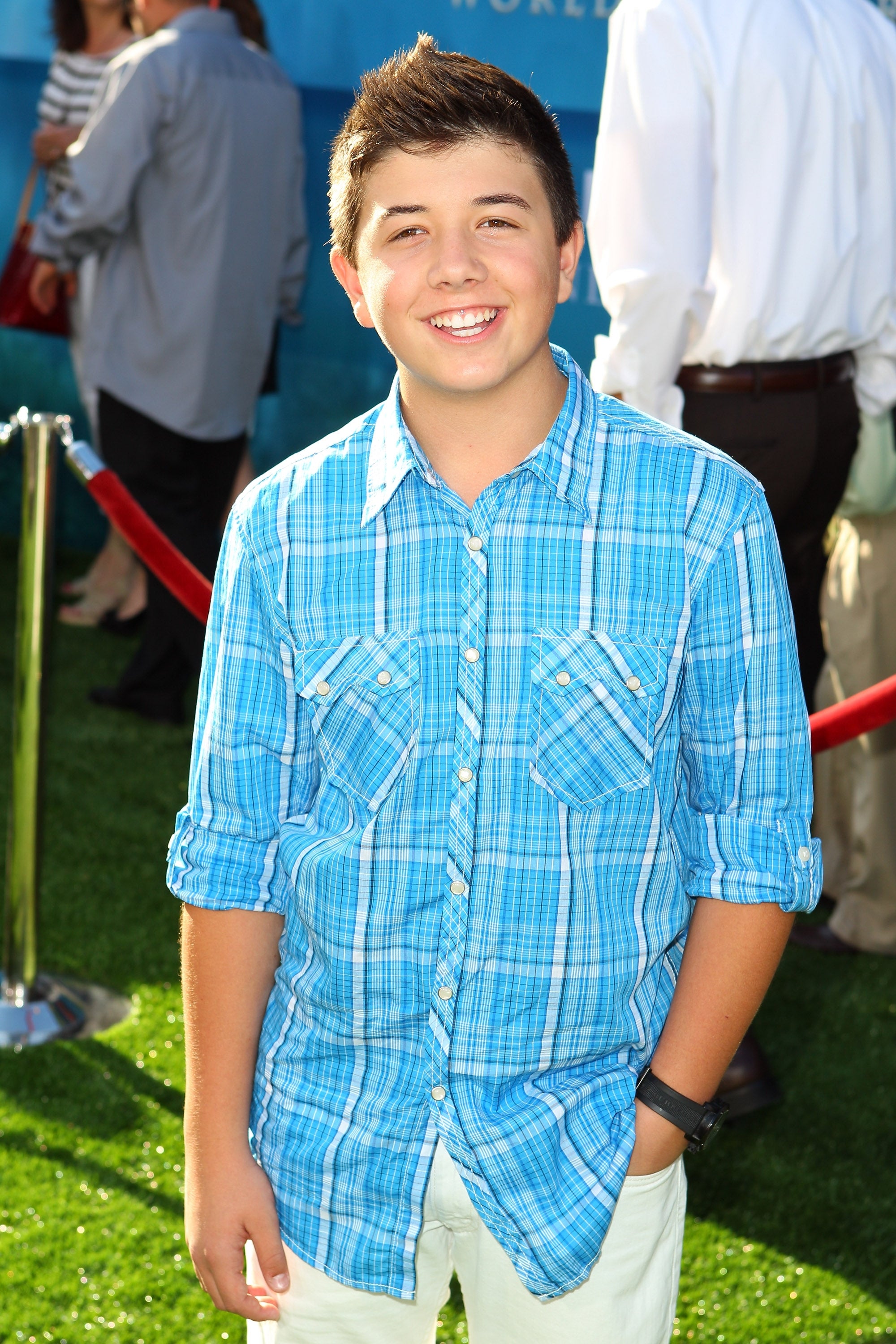 Person in a plaid shirt poses on a red carpet event with a relaxed, smiling expression