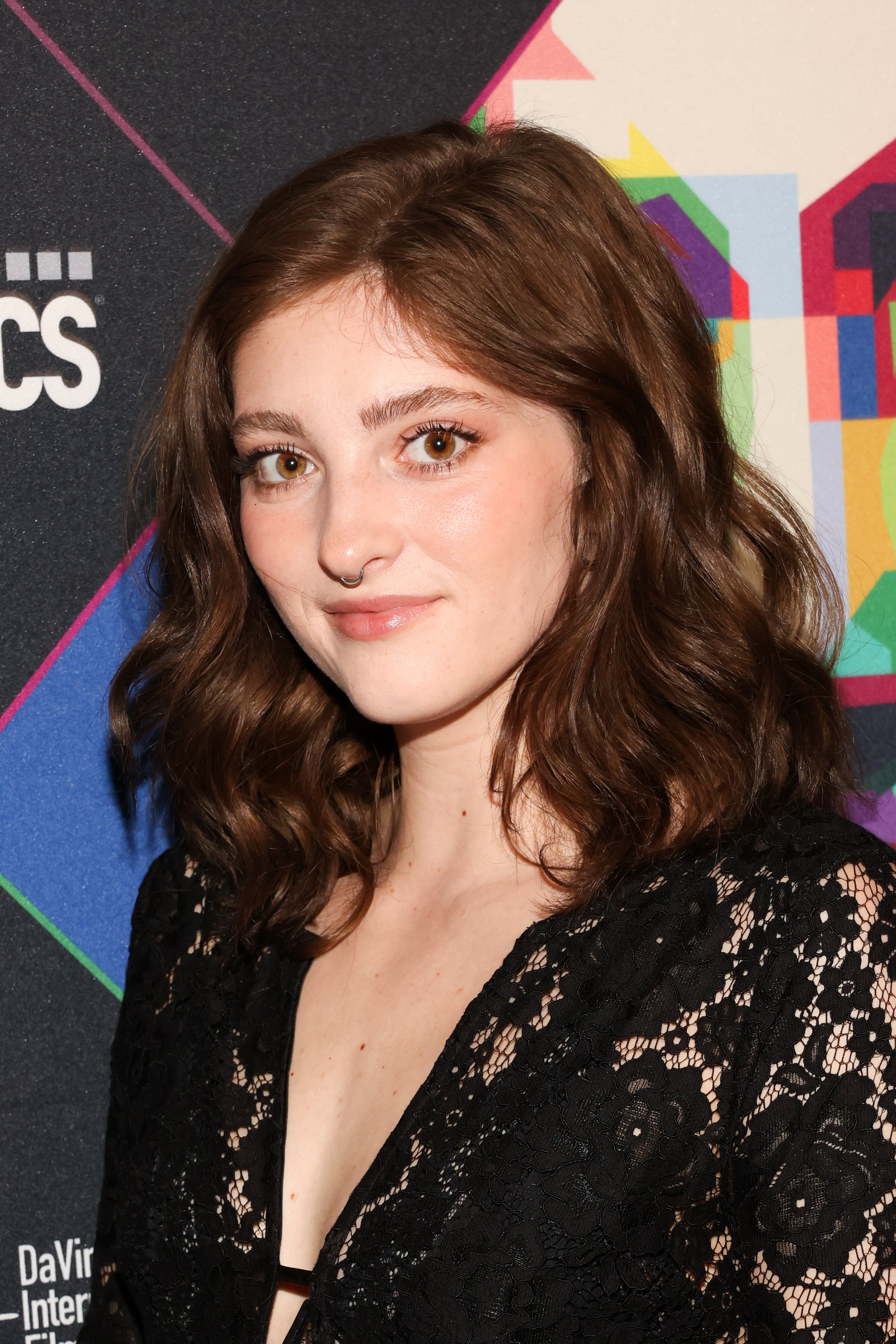 Person with shoulder-length hair in a black lace outfit stands against a patterned backdrop at a media event