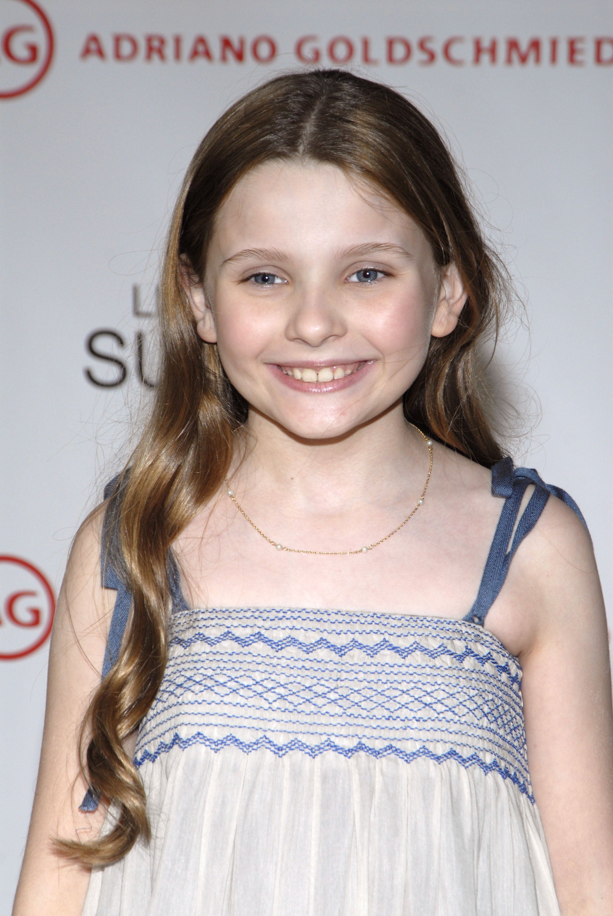 A person smiles while standing on a red carpet, wearing a sleeveless dress with a detailed upper pattern and long hair