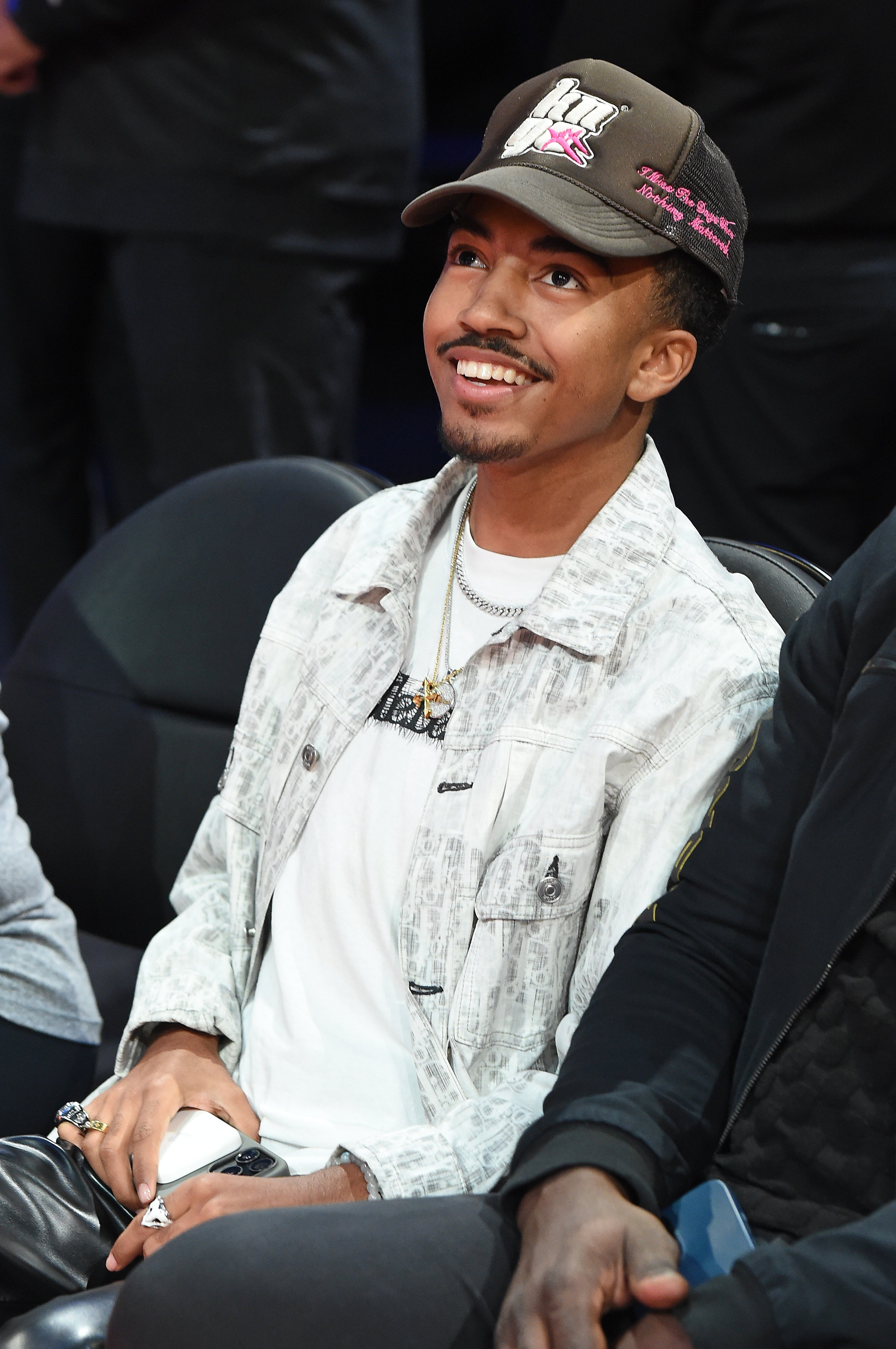 Person seated in the audience, wearing a jacket, a casual hat, and necklaces, smiling and looking upward