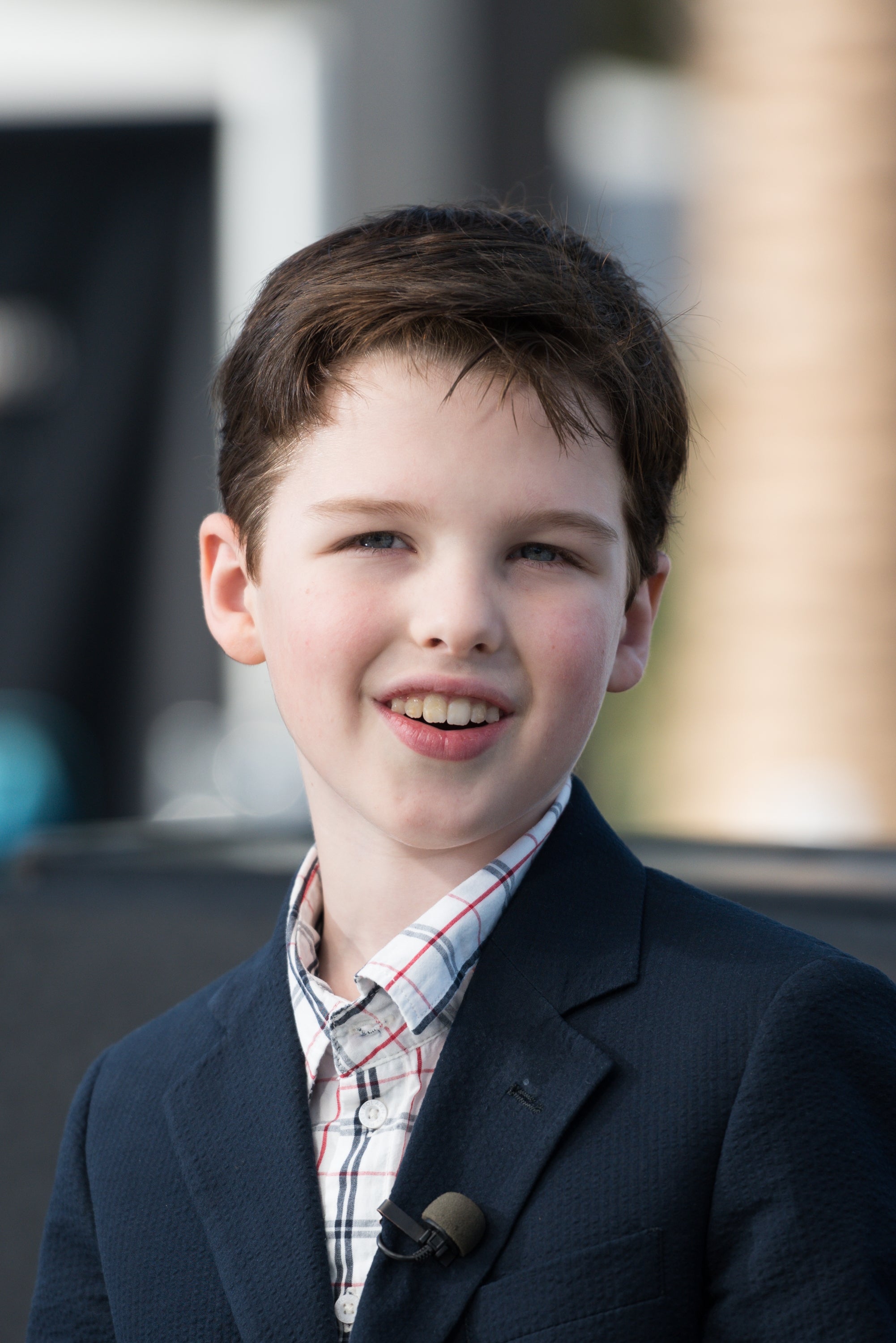 A young person in a checkered shirt and blazer smiles while being interviewed outdoors