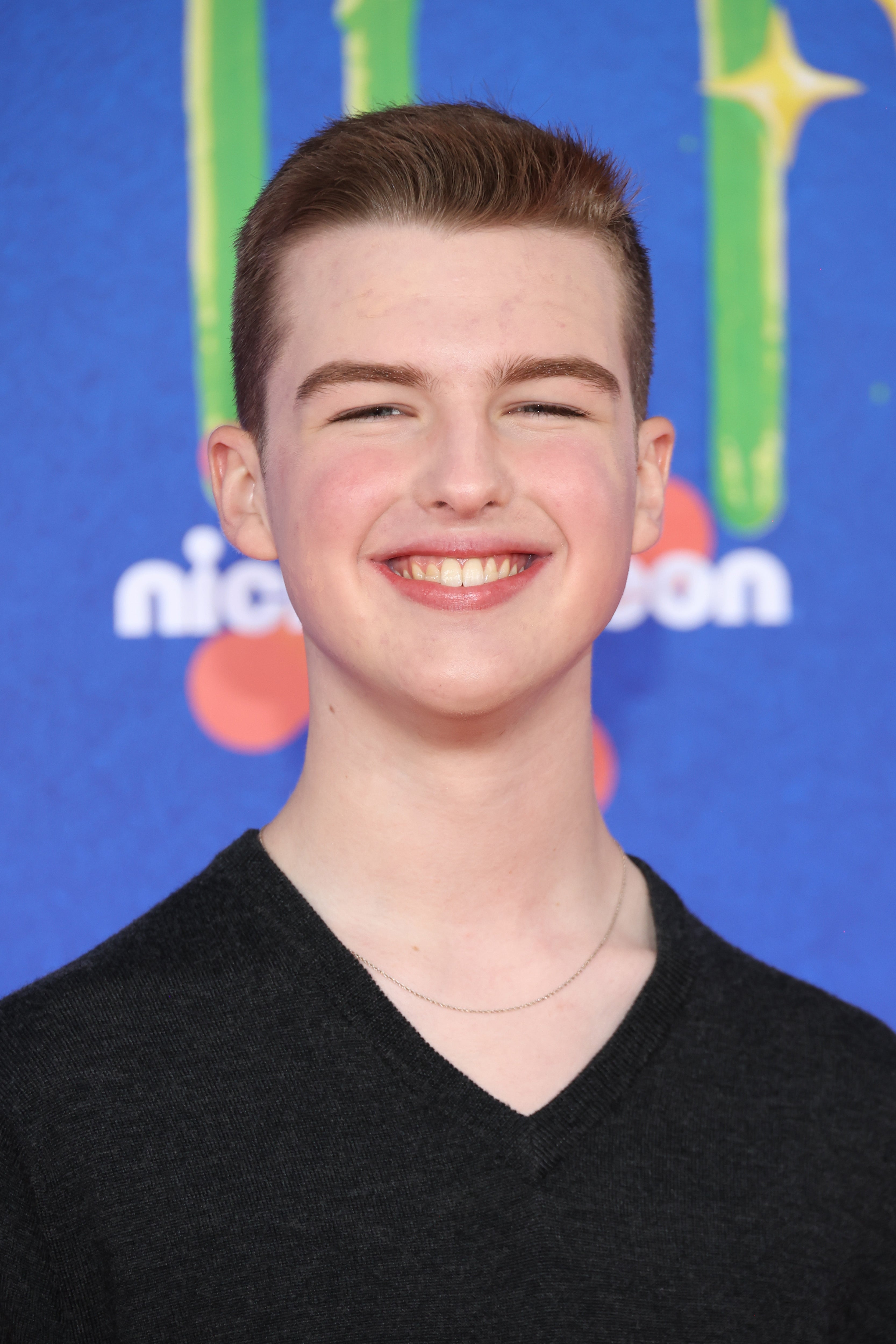 A person smiling on a red carpet wearing a simple, black V-neck shirt