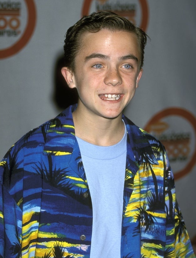 Young individual smiling on red carpet, wearing a tropical-patterned shirt over a plain shirt