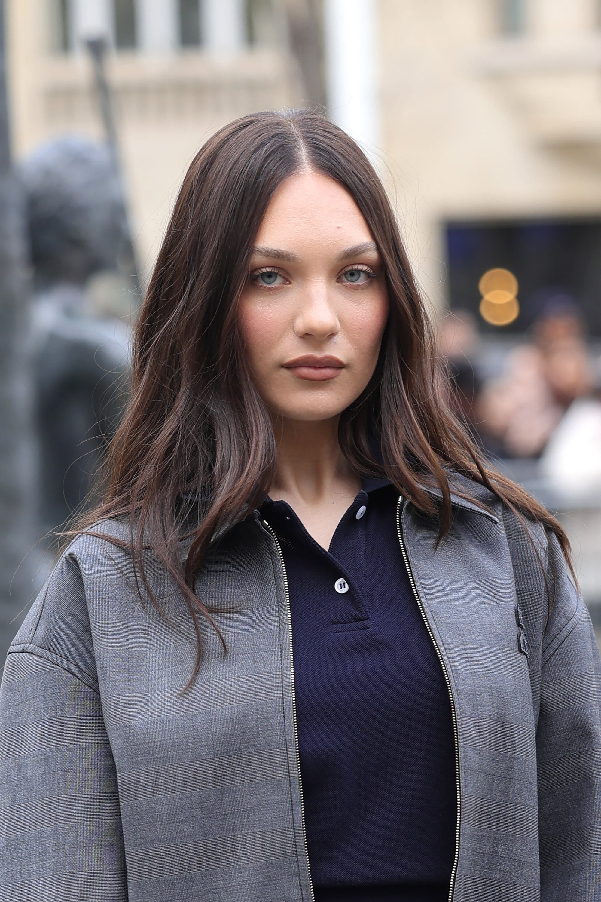 Person with long hair wearing a casual jacket over a polo shirt, standing outdoors