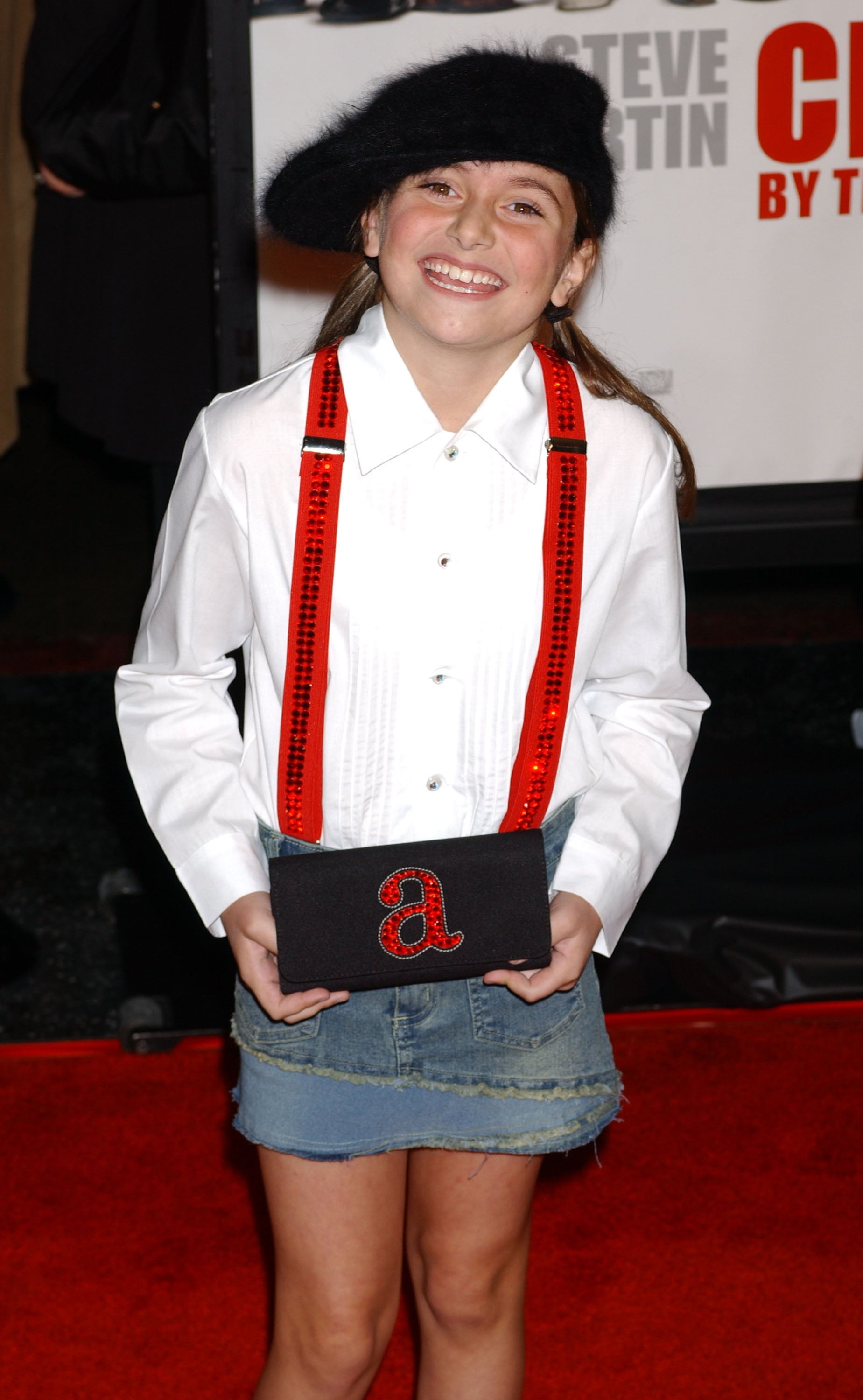 Young person on red carpet in beret, white shirt with red suspenders, denim skirt, holding a clutch