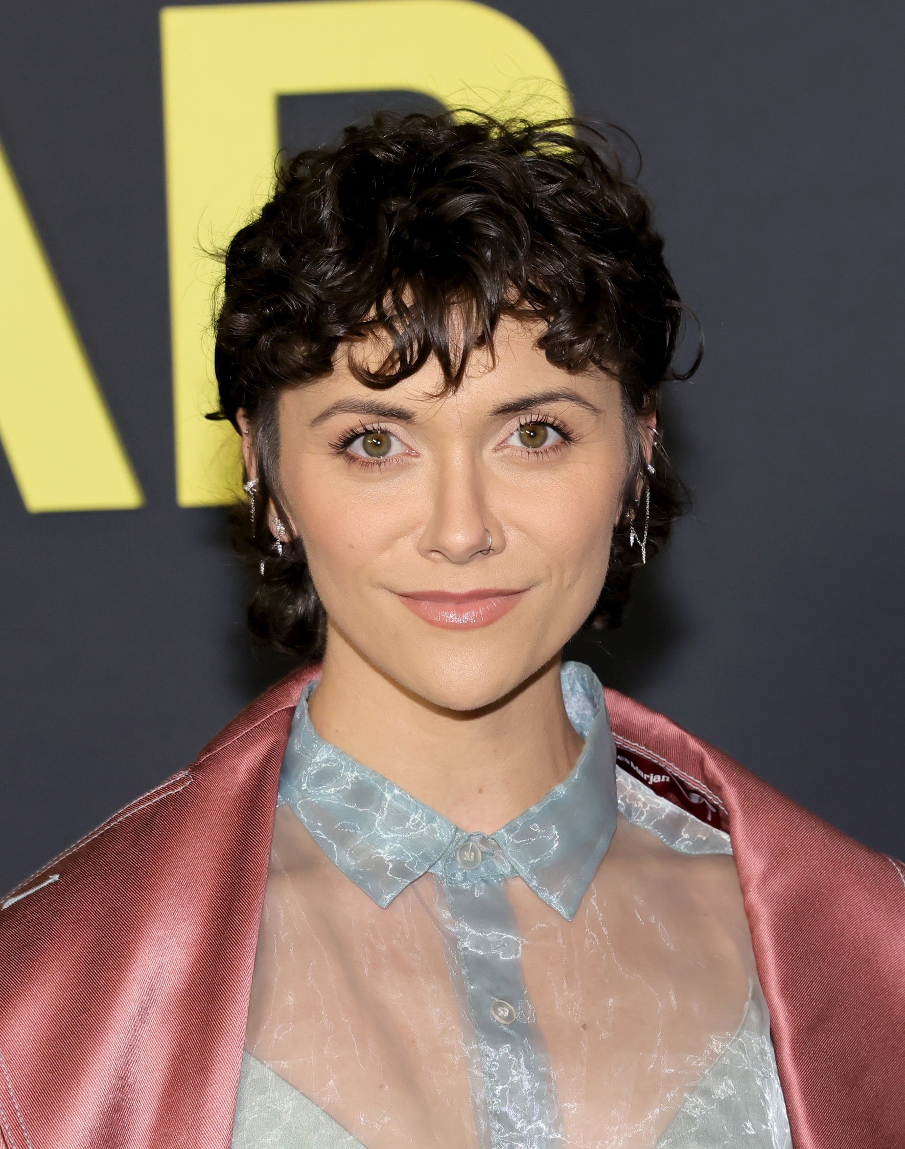 A person with short, curly hair is wearing a sheer blouse under a satin jacket at an event