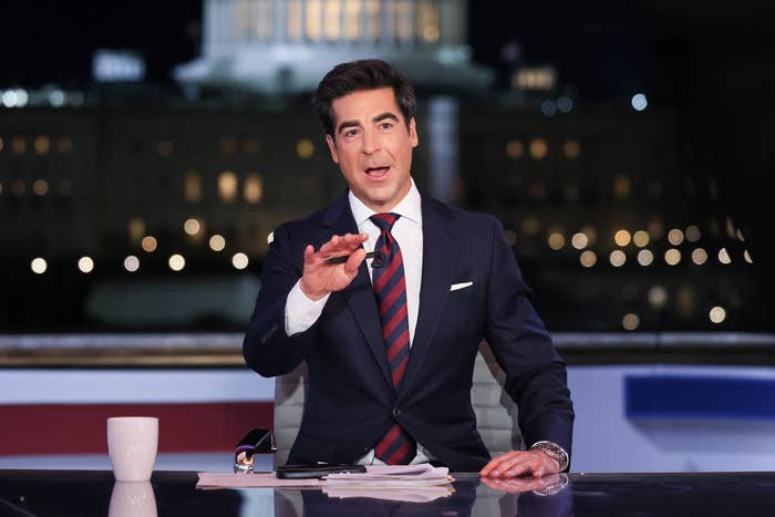 A person in a suit sits at a news desk, gesturing while speaking, with a blurred building backdrop at night
