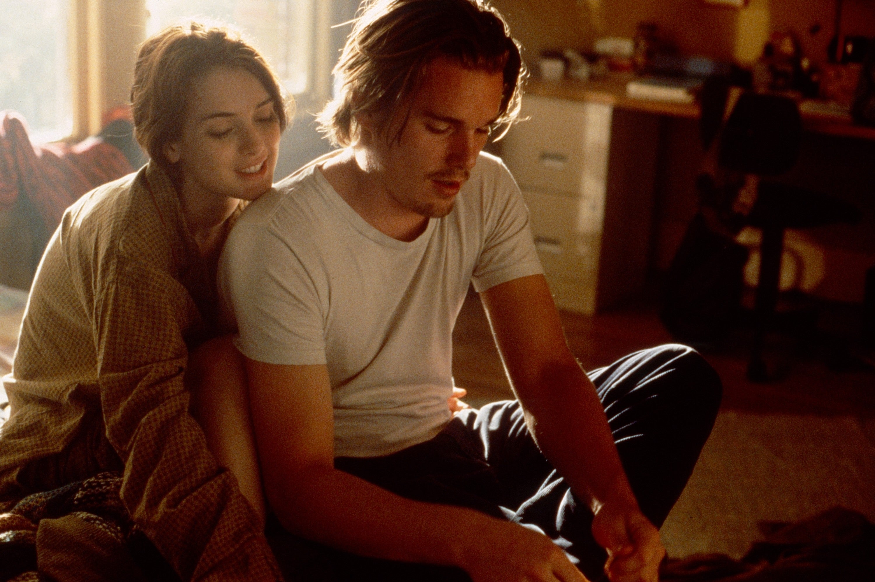A man and woman sit close together in a cozy room, sharing an intimate moment. She leans on his shoulder, both dressed in casual, relaxed clothing