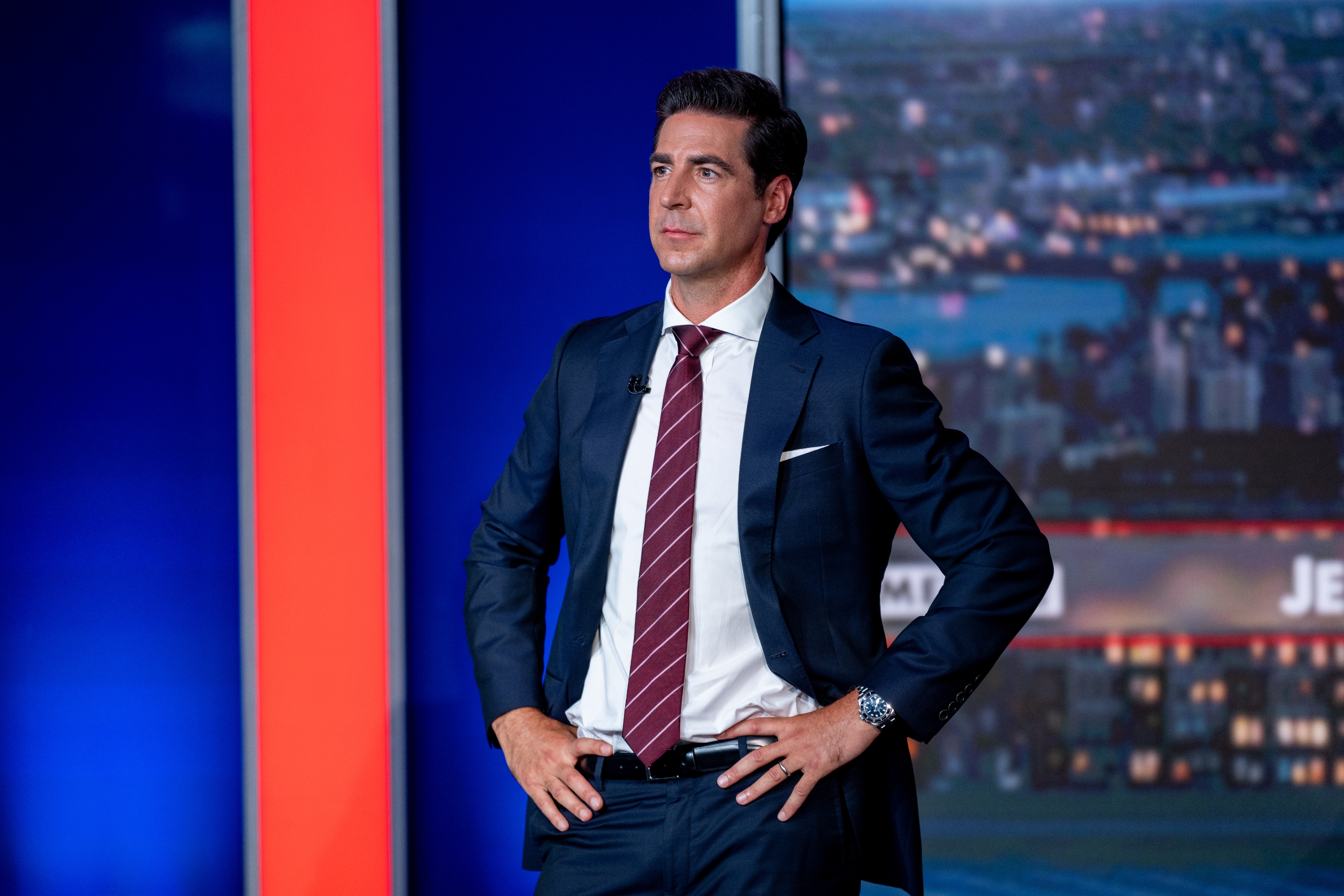 Person in a dark suit and striped tie stands confidently with hands on hips, set against a cityscape backdrop on a news set