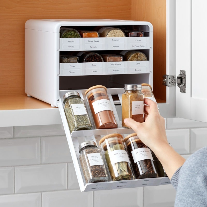 Person organizing spice jars in a tiered cabinet organizer, pulling out a jar labeled "Cumin" from a neatly arranged spice rack