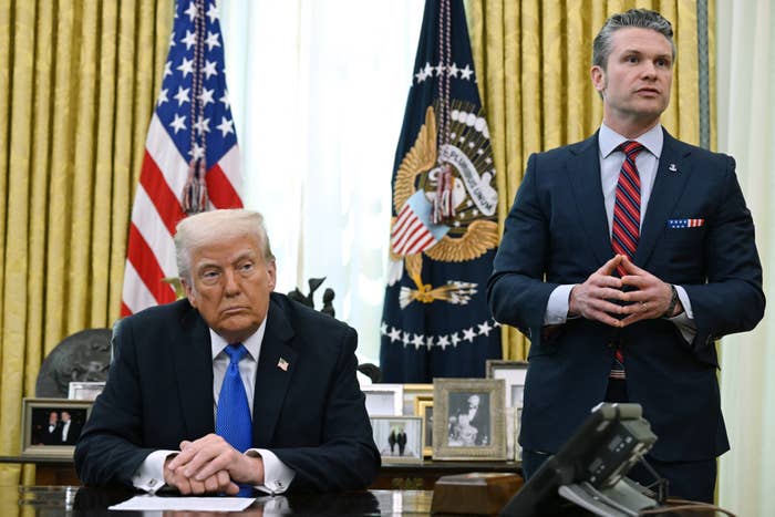 Two men in a formal setting, one seated at a desk with hands clasped, the other standing and speaking, with U.S. flags and a presidential seal in the background