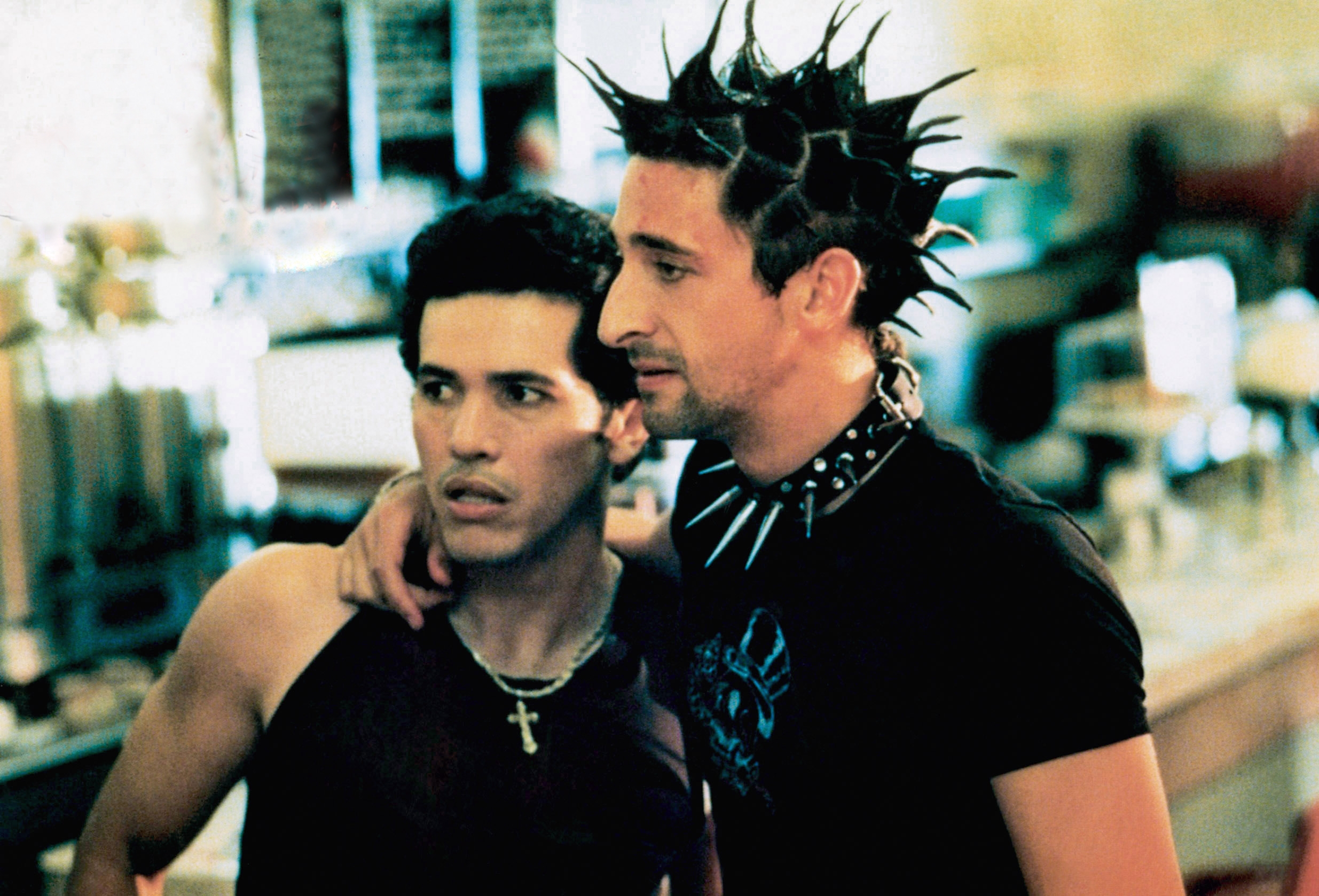 Two men stand closely, one with spiked hair and wearing a studded choker. They appear engaged, suggesting a scene from a movie or TV show