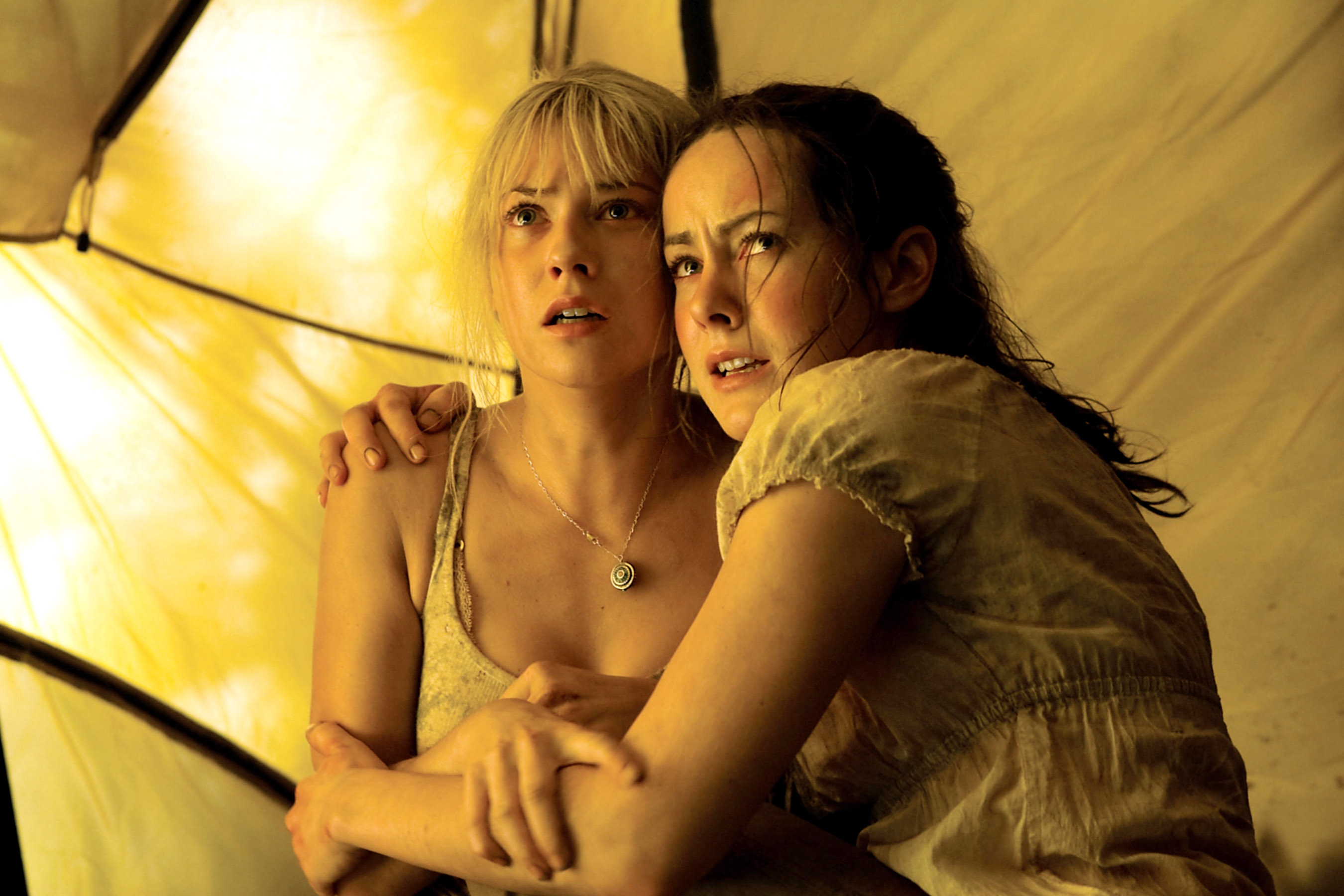 Two women in a tense scene inside a dimly lit tent, appearing anxious and holding each other