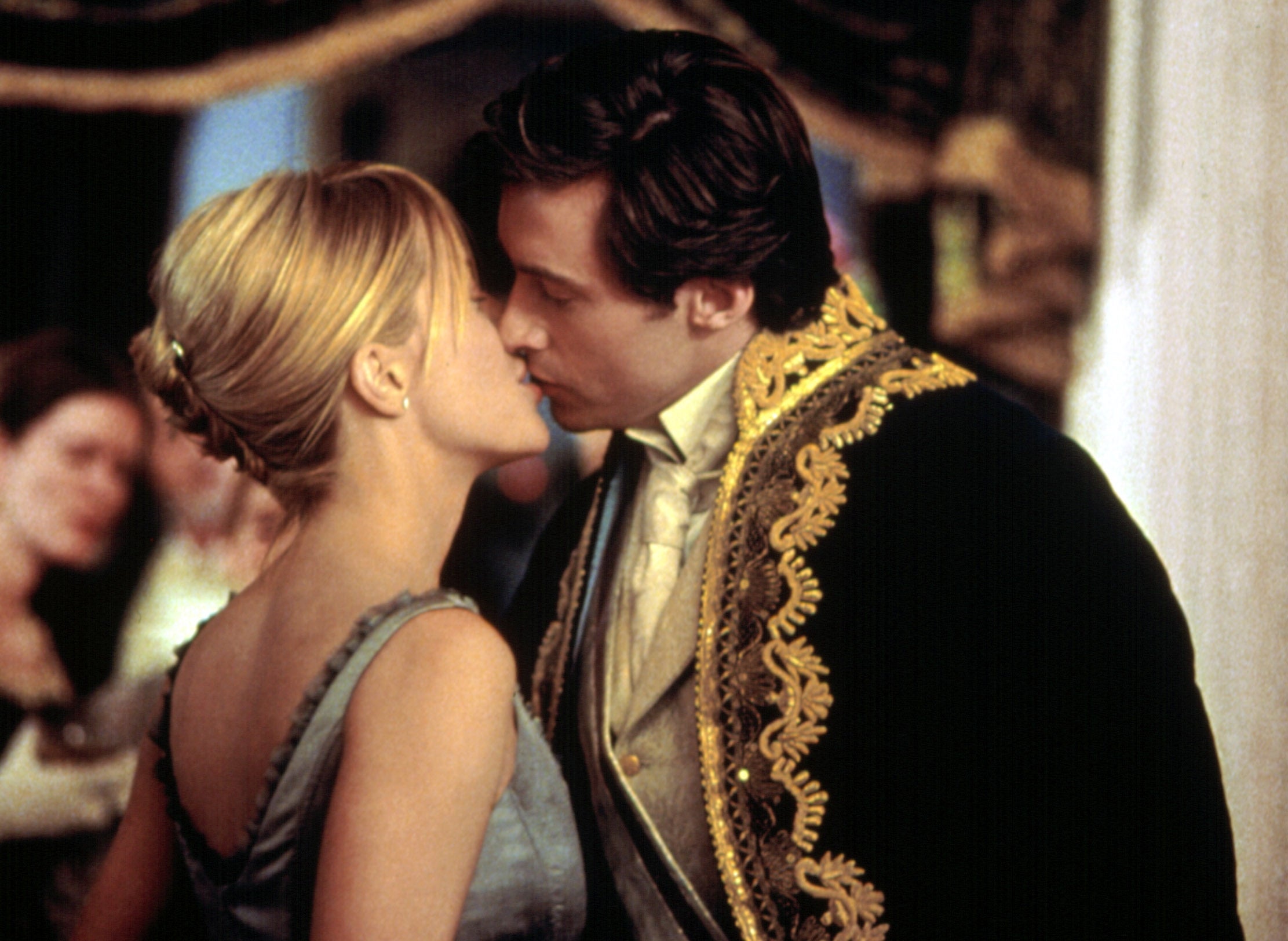 Two actors in period costumes nearly kiss at an elegant event, surrounded by onlookers. One wears an embroidered jacket, the other a simple gown