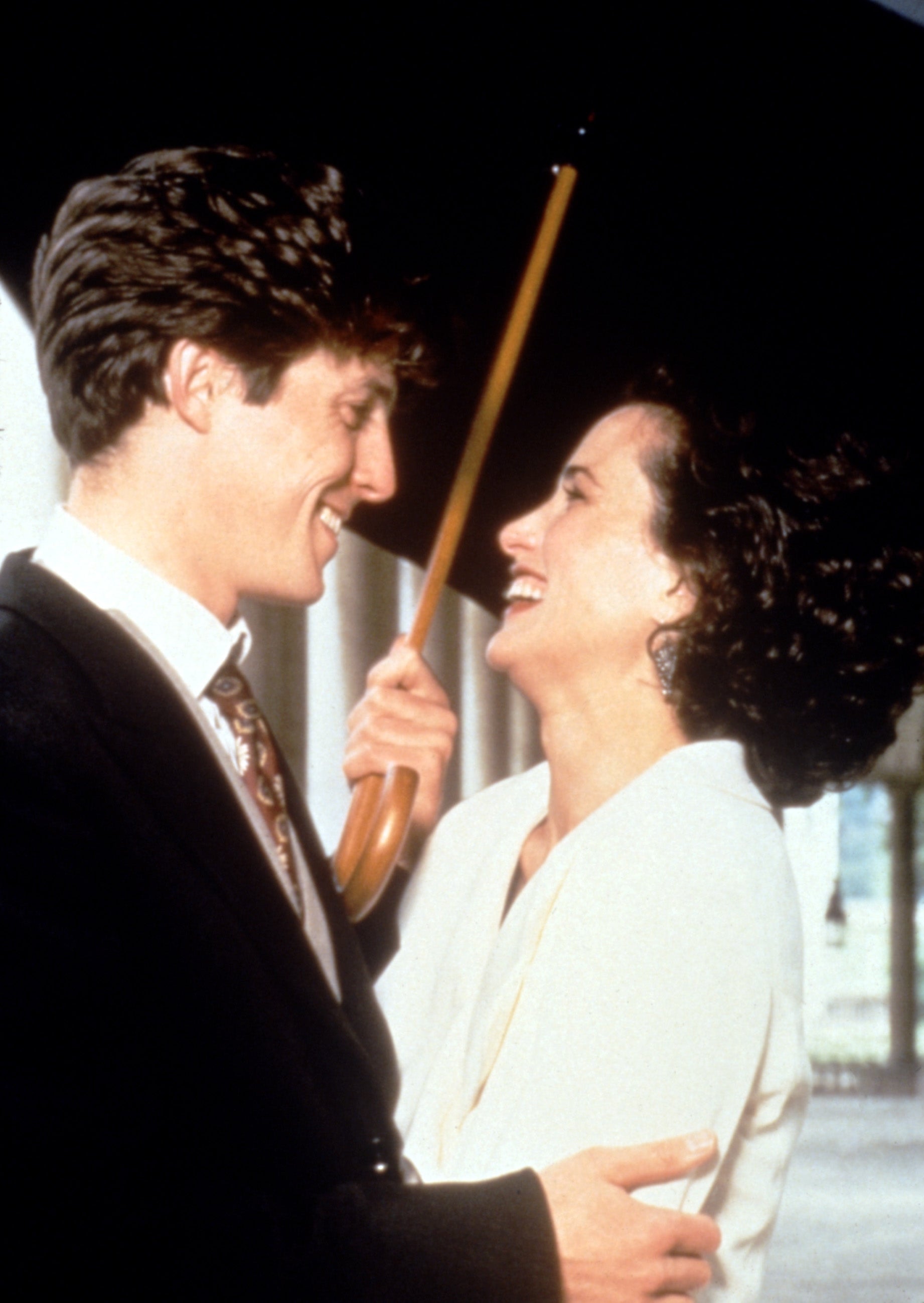 Two people smiling under an umbrella. The man wears a suit and tie; the woman in a blouse appears joyful, suggesting a lighthearted moment