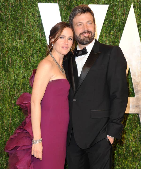 A couple poses on a red carpet; the woman wears a strapless gown with ruffled details, and the man is in a classic tuxedo