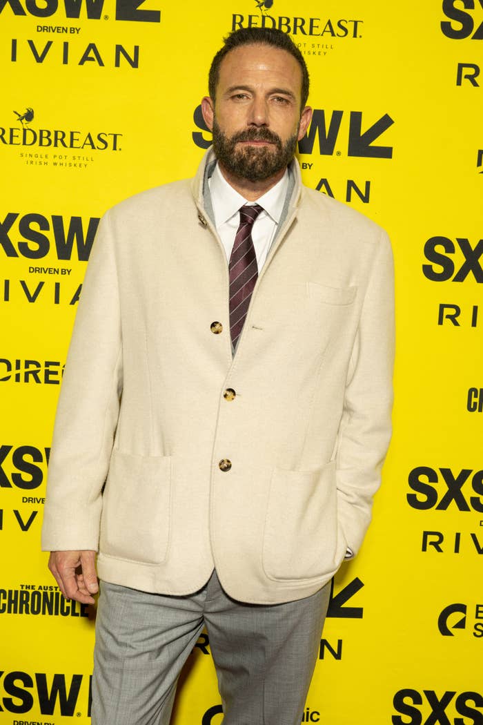 A bearded man in a light colored blazer and tie poses at a SXSW event, standing against a backdrop filled with sponsor logos