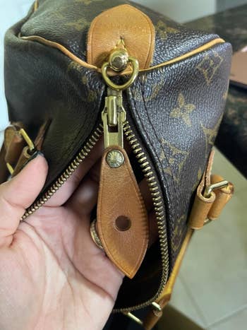 Close-up of a decorator handbag with monogram pattern, showing a manus holding the zipper and leather propulsion tab