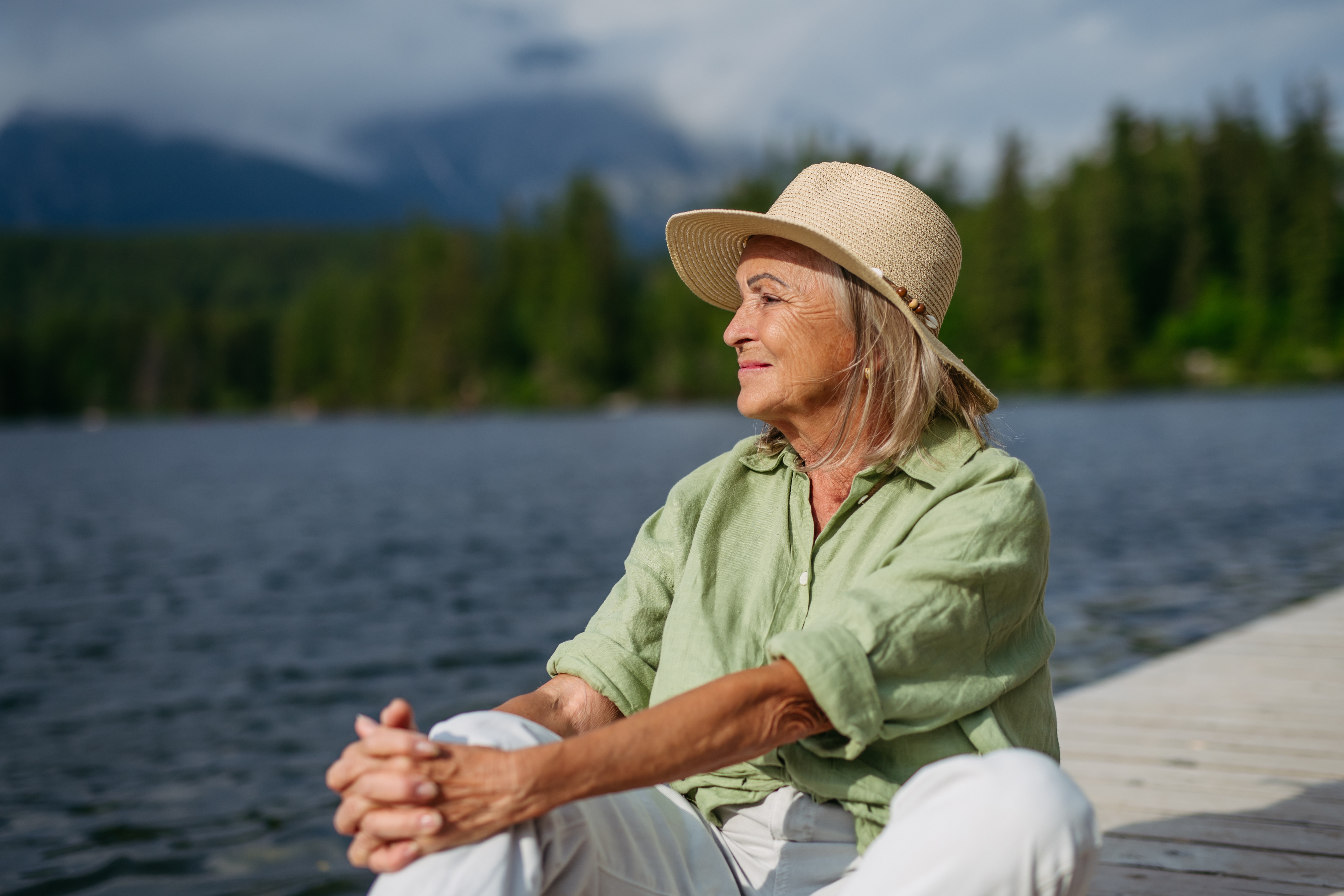 Eldre kvinne i en bredbrød hatt og uformell antrekk sitter på en brygge ved en innsjø, ser rolig og kontemplativ ut. Skog og fjell i bakgrunnen
