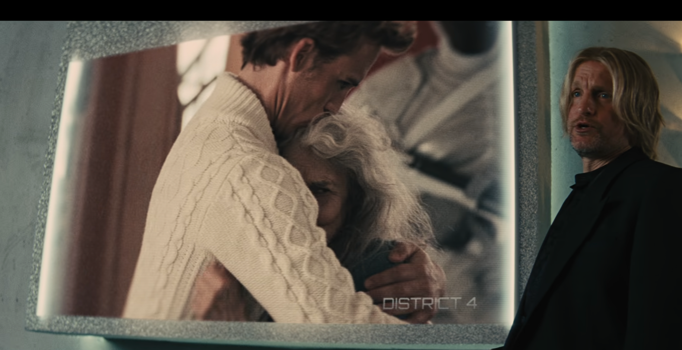 Haymitch stands beside a large screen displaying an emotional image of Finnick and Mags embracing closely in a movie scene