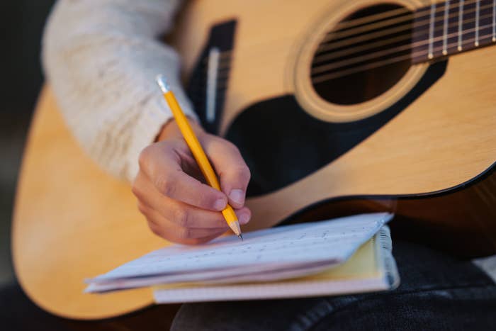 Person writing in a notebook while holding a yellow pencil, resting on an acoustic guitar