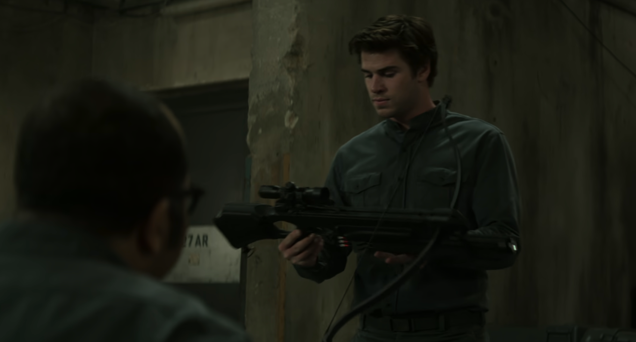 Liam Hemsworth as Gale in military-style attire, holding a weapon with Beetee nearby in a movie scene