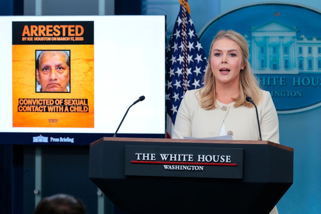 A woman at a podium speaks at a White House press briefing. Behind her is a screen displaying news of an arrest for a serious offense