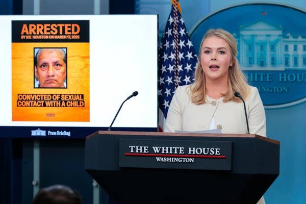A woman at a podium speaks at a White House press briefing. Behind her is a screen displaying news of an arrest for a serious offense