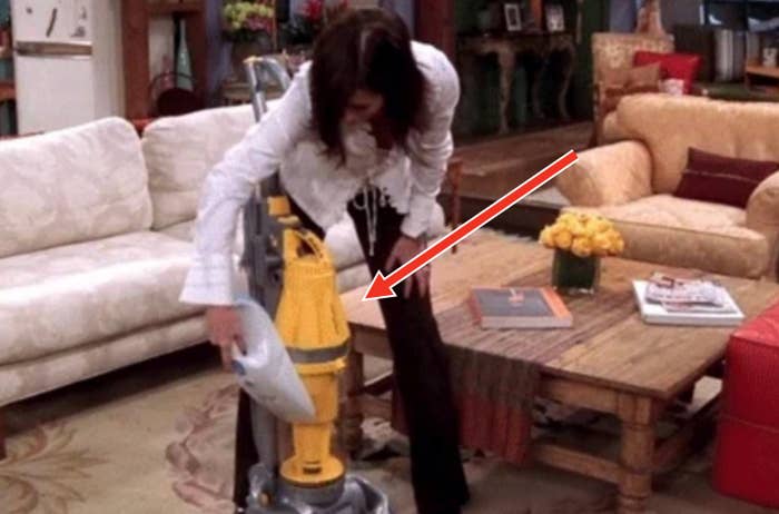 Person in a cozy living room bending down to use a vacuum cleaner near a coffee table with books and a vase of yellow flowers