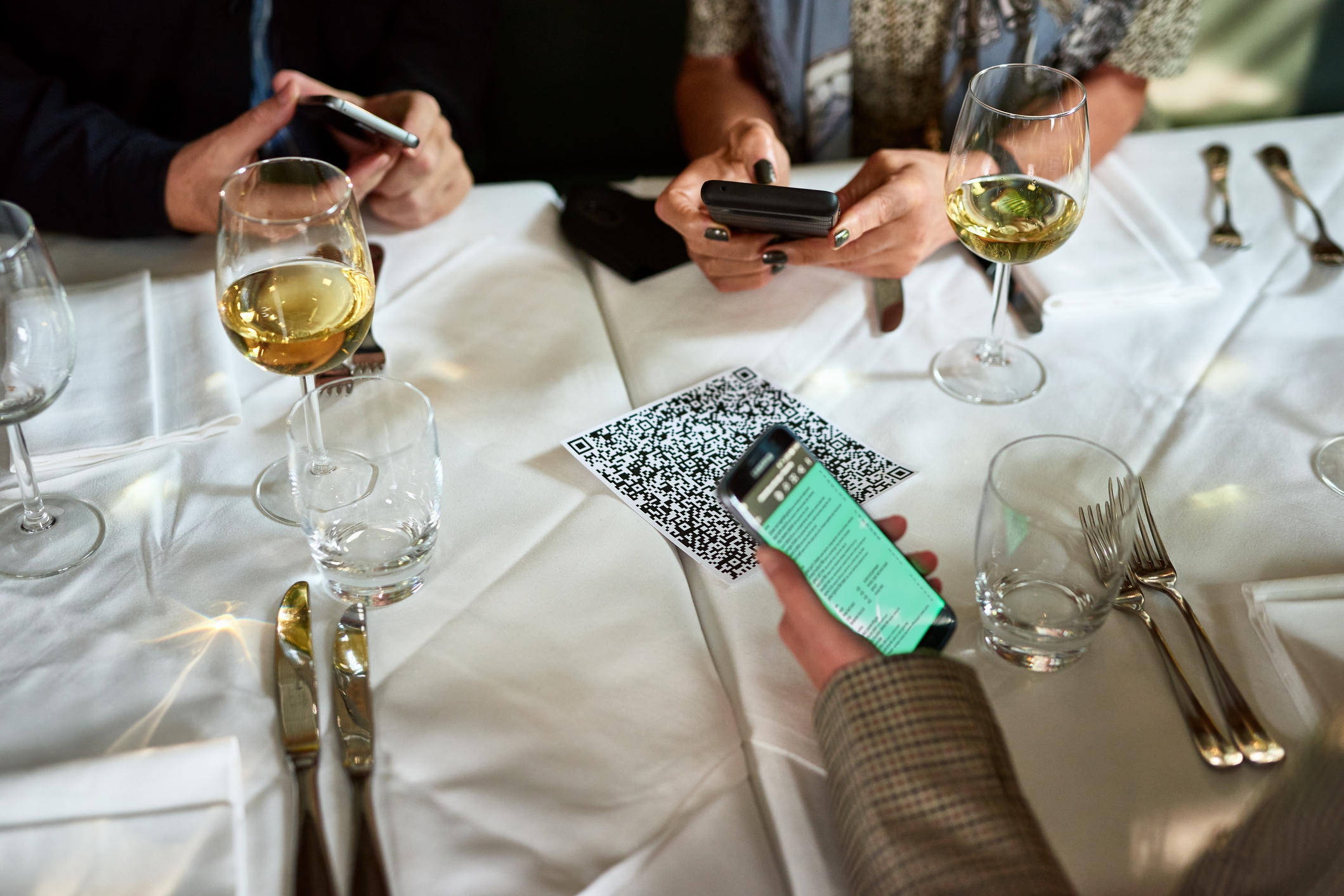 Tre personer ved et bord ved hjelp av smarttelefoner for å skanne en QR -kode, med vinglass i nærheten