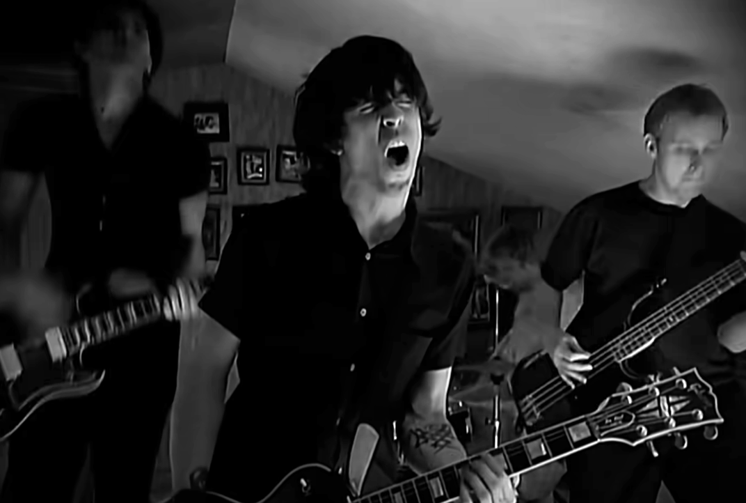 Band playing instruments with intensity in a dimly lit room; lead singer in center, expressive in delivery