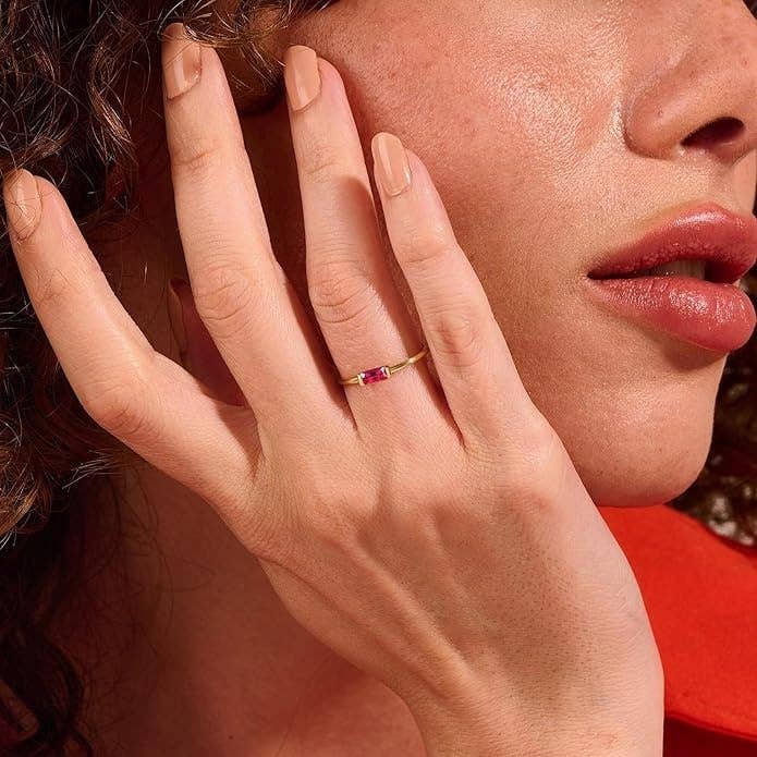 Person wearing a thin gold ring with a small red gemstone, touching their face, highlighting the jewelry for a shopping feature