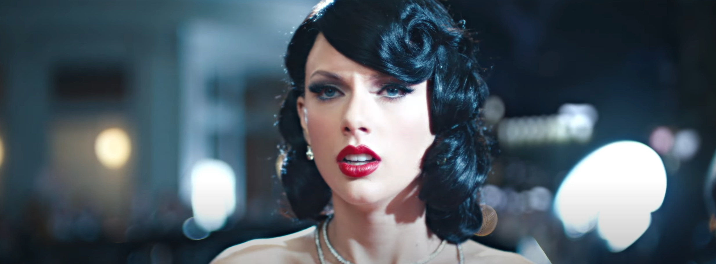 Person with black, wavy hair and bright red lips, wearing a diamond necklace, looks surprised in a glamorous, possibly event-like setting