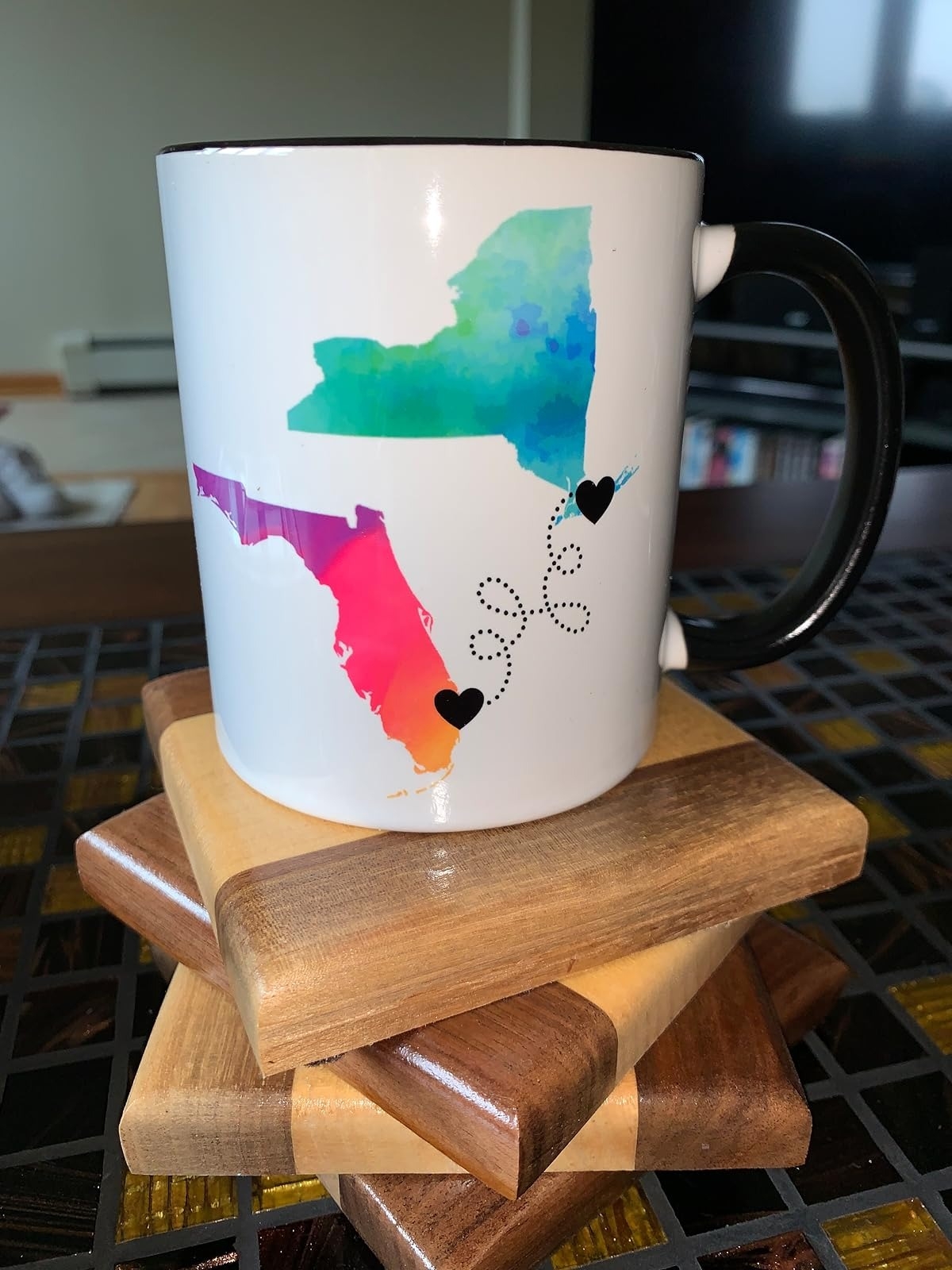 Mug with colorful map outlines of New York and Florida, connected by a dotted line with heart icons, placed on wooden coasters