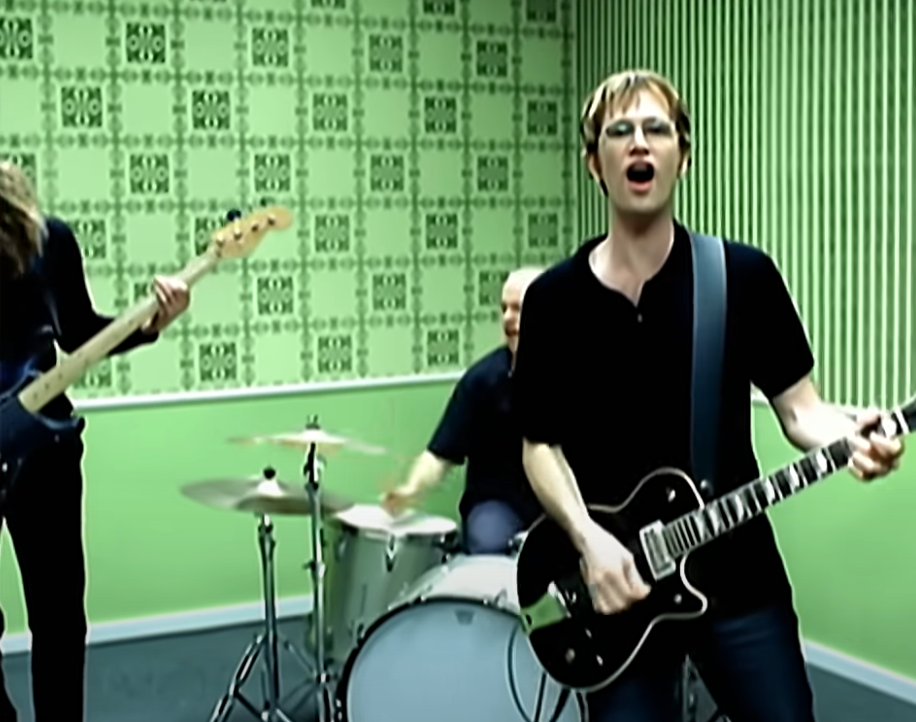 Band performing in a room with patterned wallpaper; guitarist and drummer visible, capturing a rock music vibe