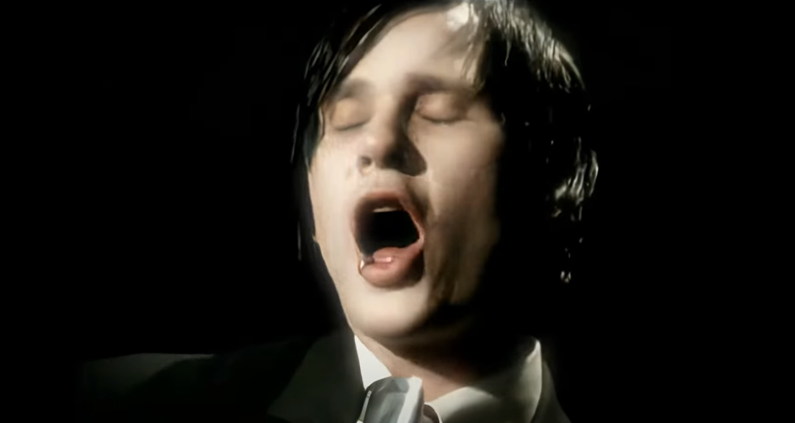 Person in a dark setting, wearing a suit, with eyes closed, passionately singing or speaking into a microphone
