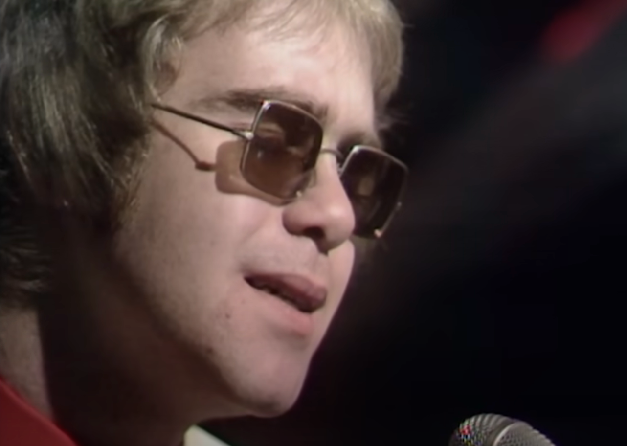 A person wearing sunglasses sings into a microphone during a performance