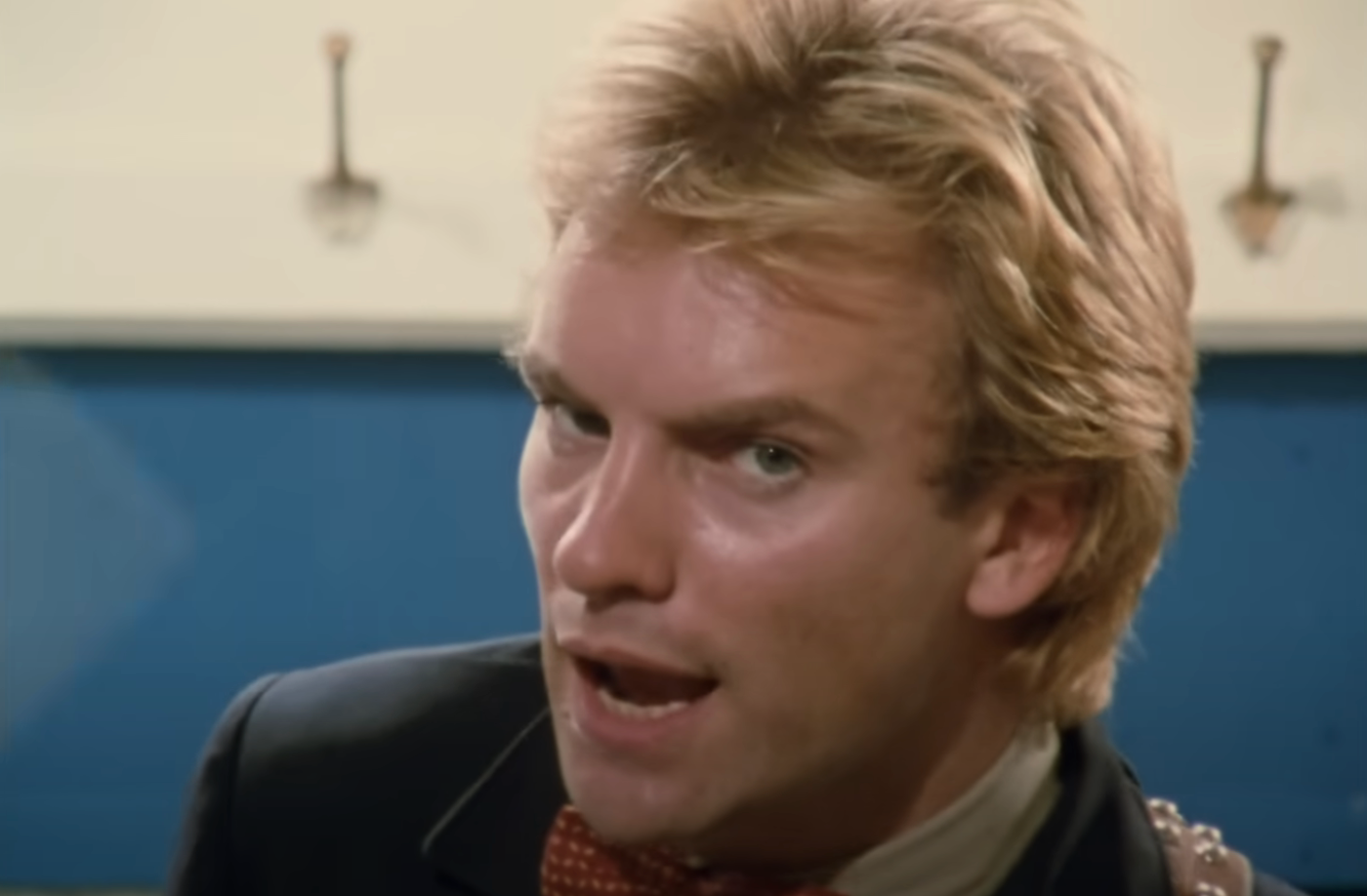 A person with short hair, wearing a jacket and bow tie, sings in a close-up. Background is slightly blurred