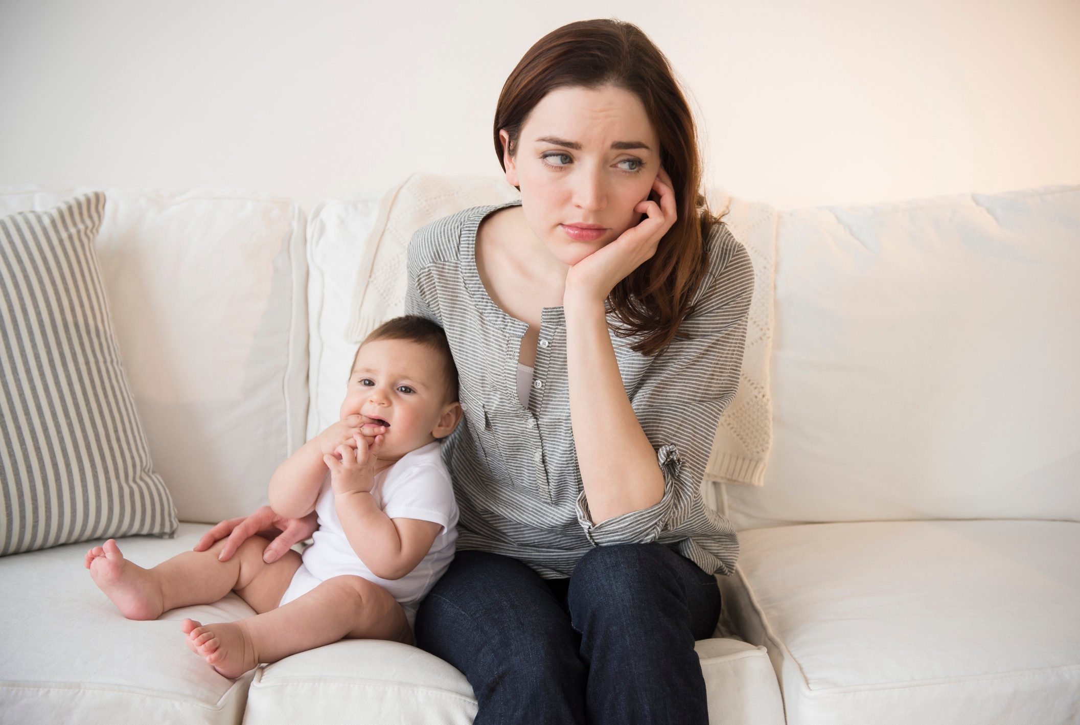 Kvinne som ser ettertenksom ut mens hun sitter på en sofa ved siden av en baby som leker med tærne