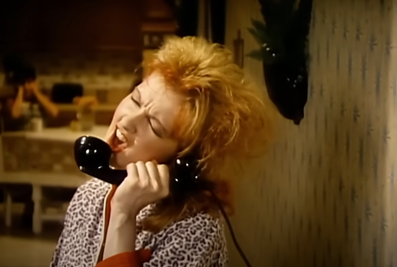 Person singing passionately into a vintage phone, wearing a patterned top, in a domestic setting
